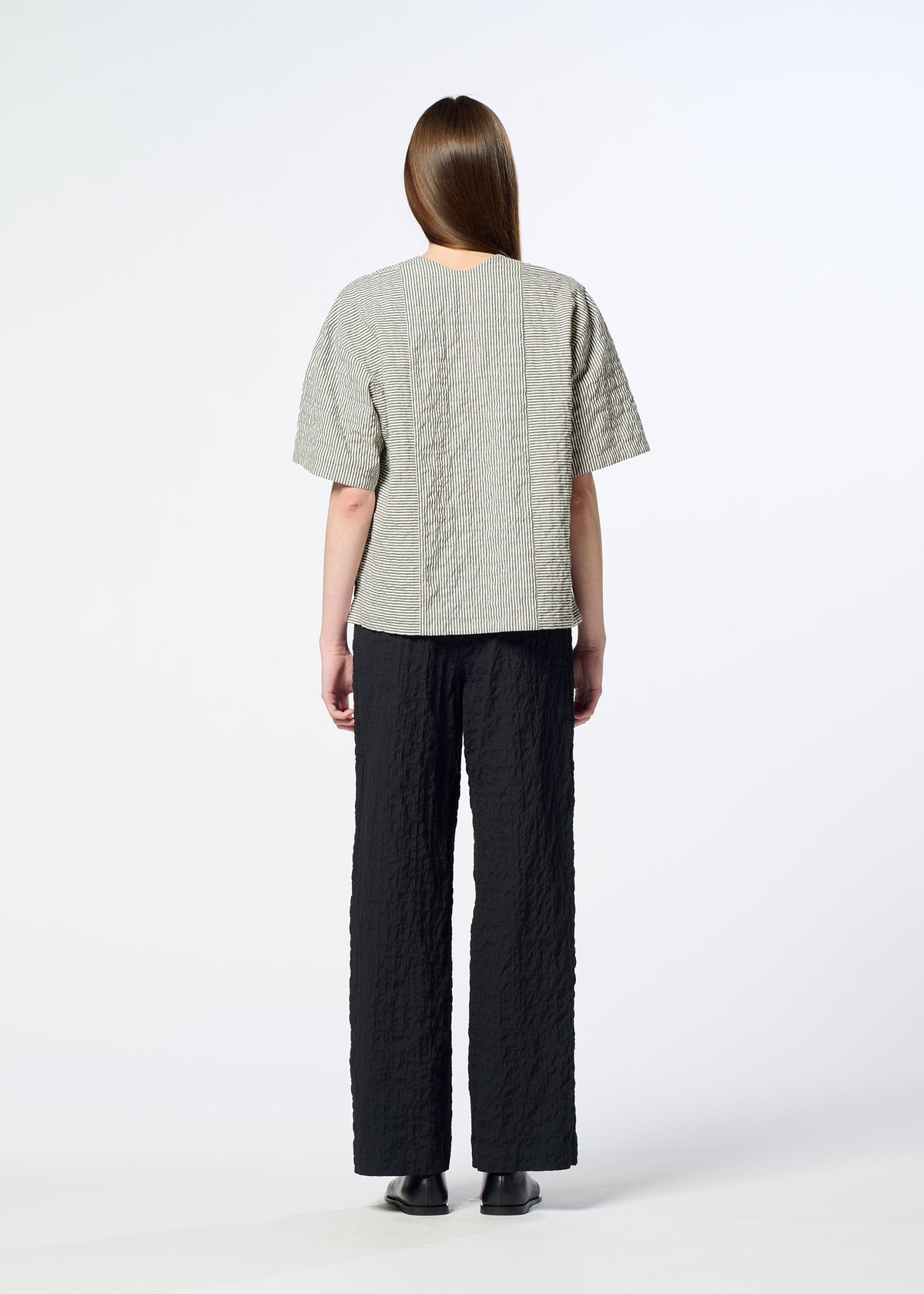 A person with long brown hair stands facing away against a plain white background, wearing HaaT’s RIPPLE STRIPE textured gray seersucker top with short sleeves, paired with black textured pants and black shoes. view 7 of 7
