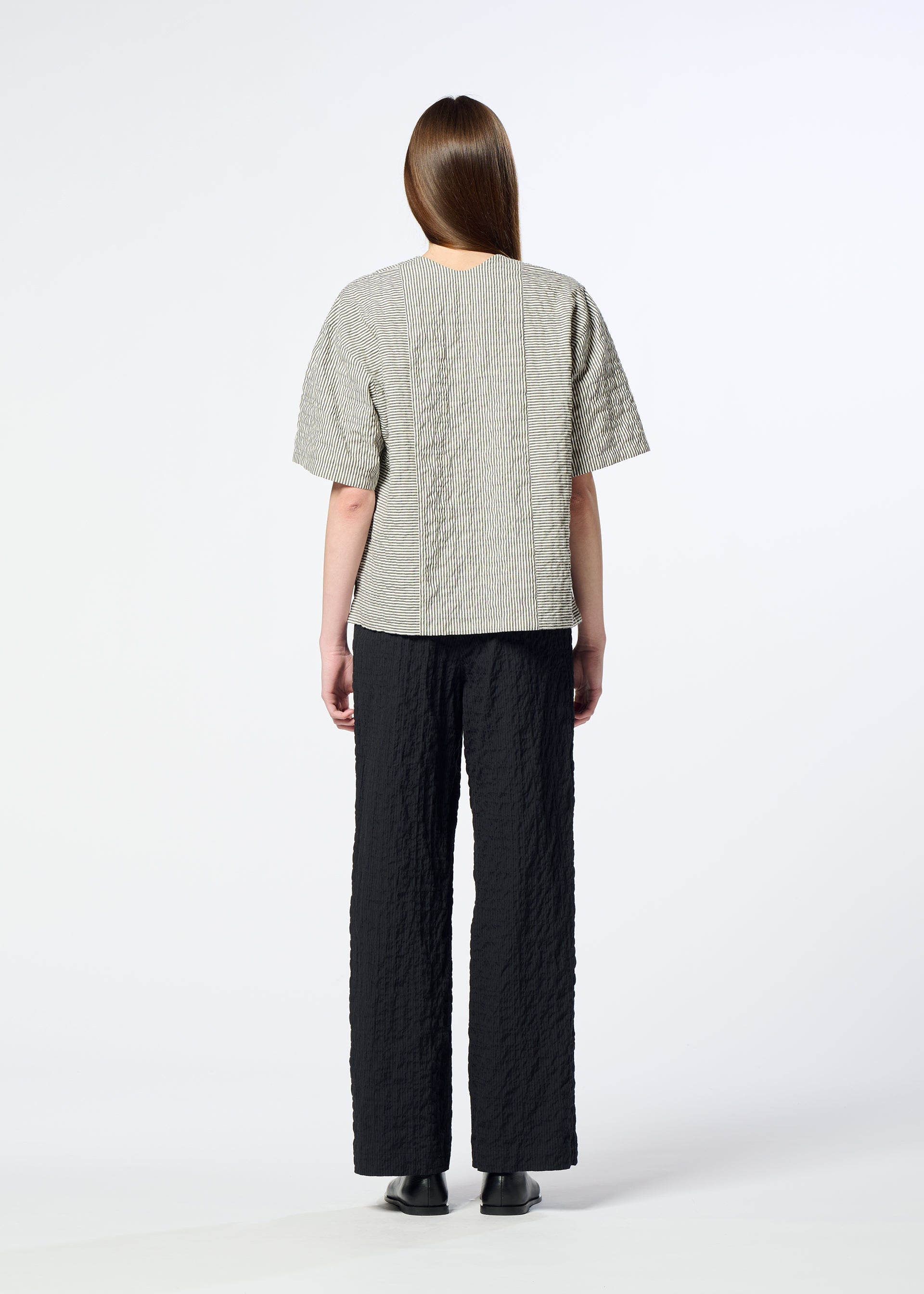 Zoomed view A person with long brown hair stands facing away against a plain white background, wearing HaaT’s RIPPLE STRIPE textured gray seersucker top with short sleeves, paired with black textured pants and black shoes.