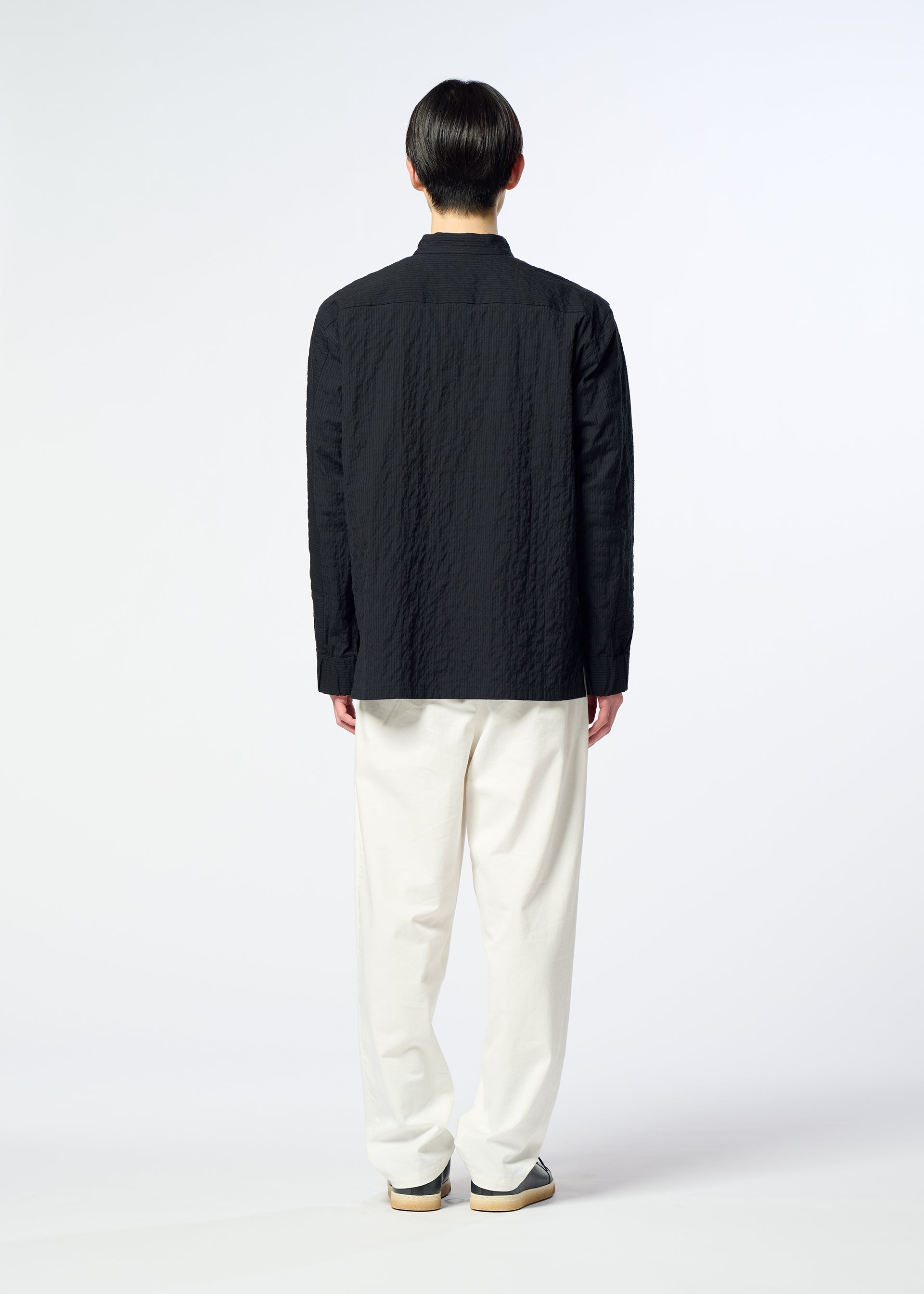 Zoomed view A person stands facing away, wearing HaaT’s RIPPLE STRIPE textured black seersucker shirt with long sleeves, paired with white loose pants and sneakers, set against a plain light background.