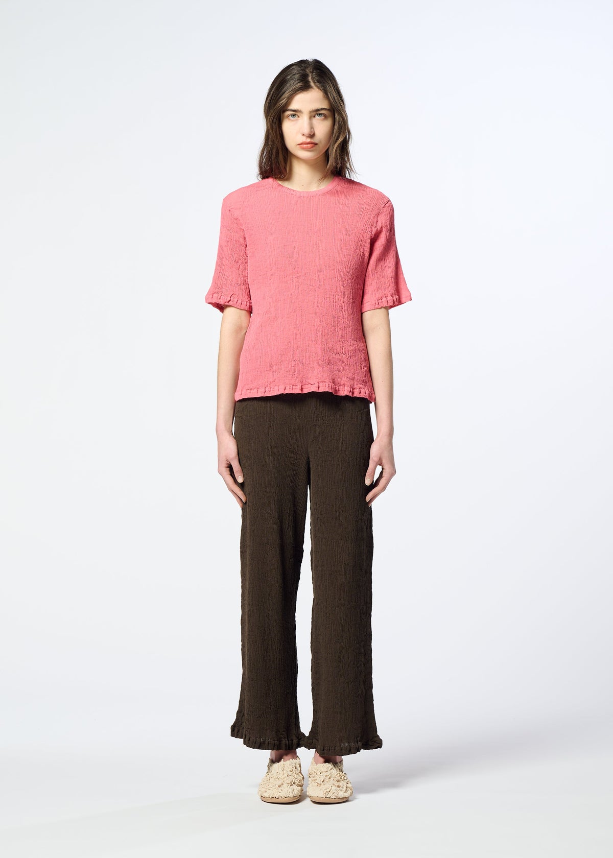 A person stands against a plain white background wearing the HaaT KYO CHIJIMI AUGUST short-sleeve top, crafted from ultra-fine cotton with a subtle crinkled texture, paired with wide-leg brown pants and beige shoes. view 5 of 7