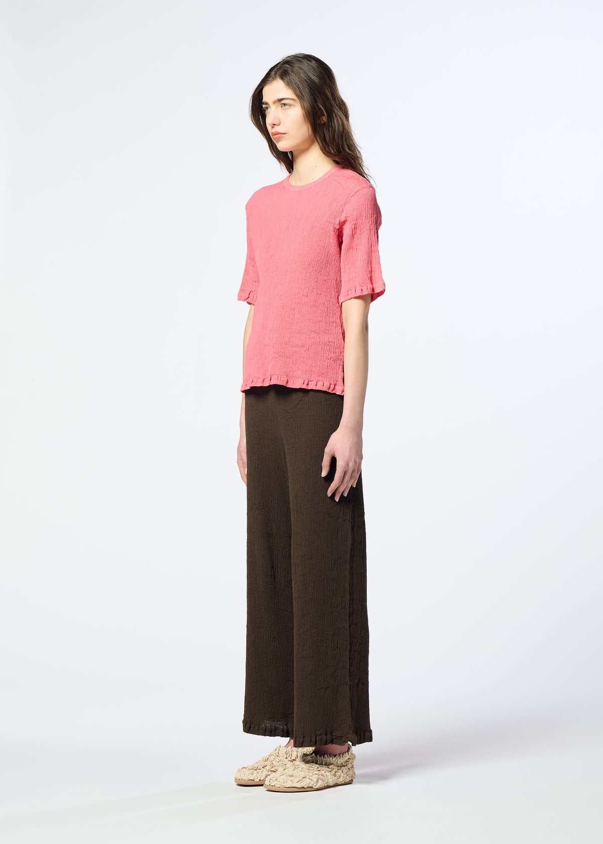 A woman stands sideways against a plain light background, wearing the HaaT KYO CHIJIMI AUGUST—a textured pink short-sleeve top crafted from ultra-fine cotton—paired with loose dark brown wide-leg pants and beige woven shoes. view 6 of 7