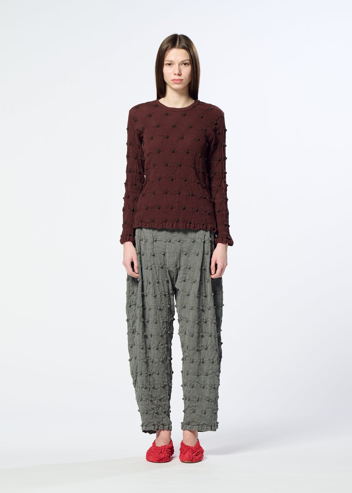 A woman stands facing forward against a plain white background, wearing the HaaT KYO CHIJIMI SQUARE EMB embroidered long-sleeved top in maroon, paired with loose gray textured pants and red shoes. view 6 of 8