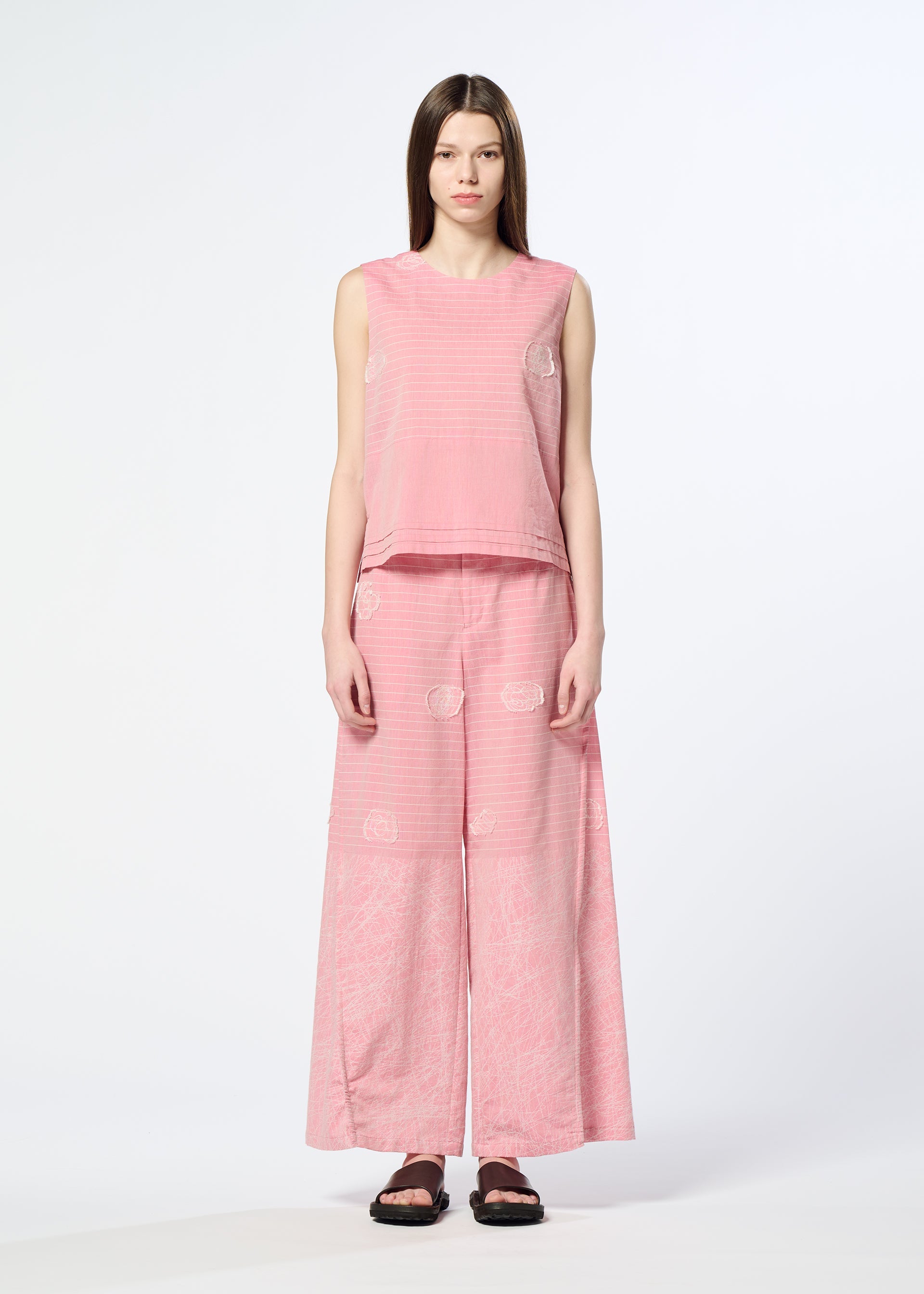 Zoomed view A woman with straight brown hair wears HaaT's ONE STROKE DRAW jacquard sleeveless top and matching wide-leg pants in a cotton-cupra blend, featuring subtle three-dimensional patterns. She pairs them with black sandals against a white background.