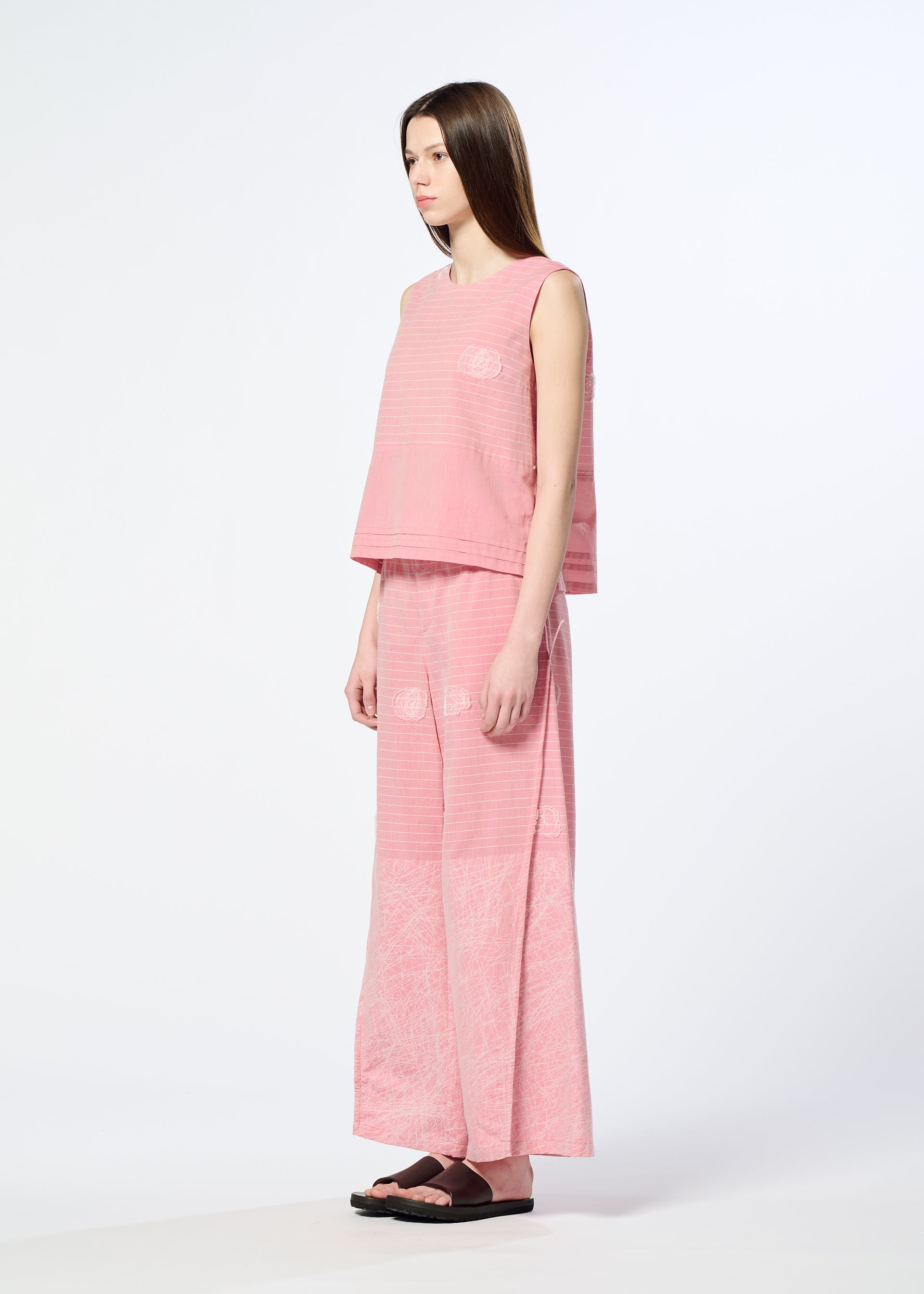 Zoomed view A woman with long brown hair and black sandals stands sideways against a plain light background, wearing HaaT’s ONE STROKE DRAW: a loose-fitting jacquard cotton-cupra blend sleeveless top and matching wide-leg pants with subtle patterns.