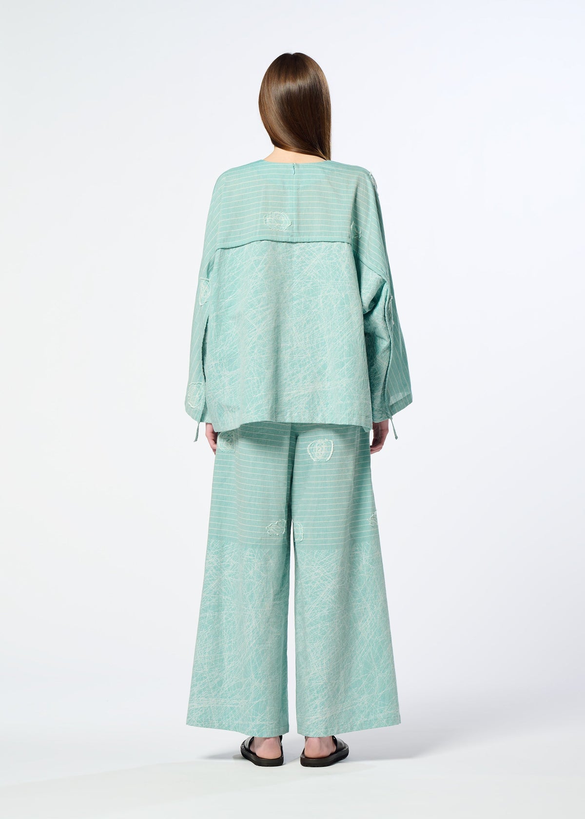 A person with long brown hair, seen from behind, wears HaaT's ONE STROKE DRAW—a loose light teal jacquard cotton cupra top and matching wide-leg pants—paired with black flat sandals, set against a plain white background. view 10 of 10
