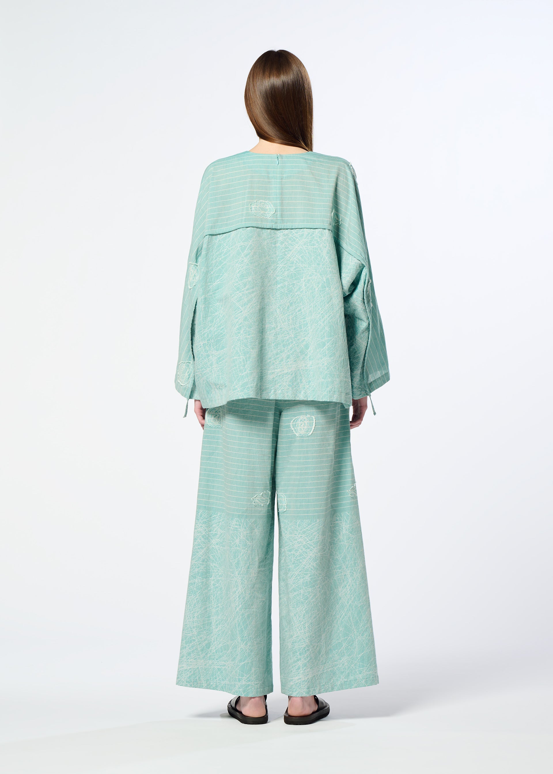 Zoomed view A person with long brown hair, seen from behind, wears HaaT's ONE STROKE DRAW—a loose light teal jacquard cotton cupra top and matching wide-leg pants—paired with black flat sandals, set against a plain white background.