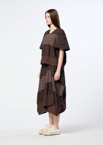 A woman stands in profile against a plain light background, wearing HaaT’s COLLAGE STRETCH Ⅱ—a relaxed, loose brown dress with horizontal beige stripes crafted from stretchy yarn—paired with white sneakers.
