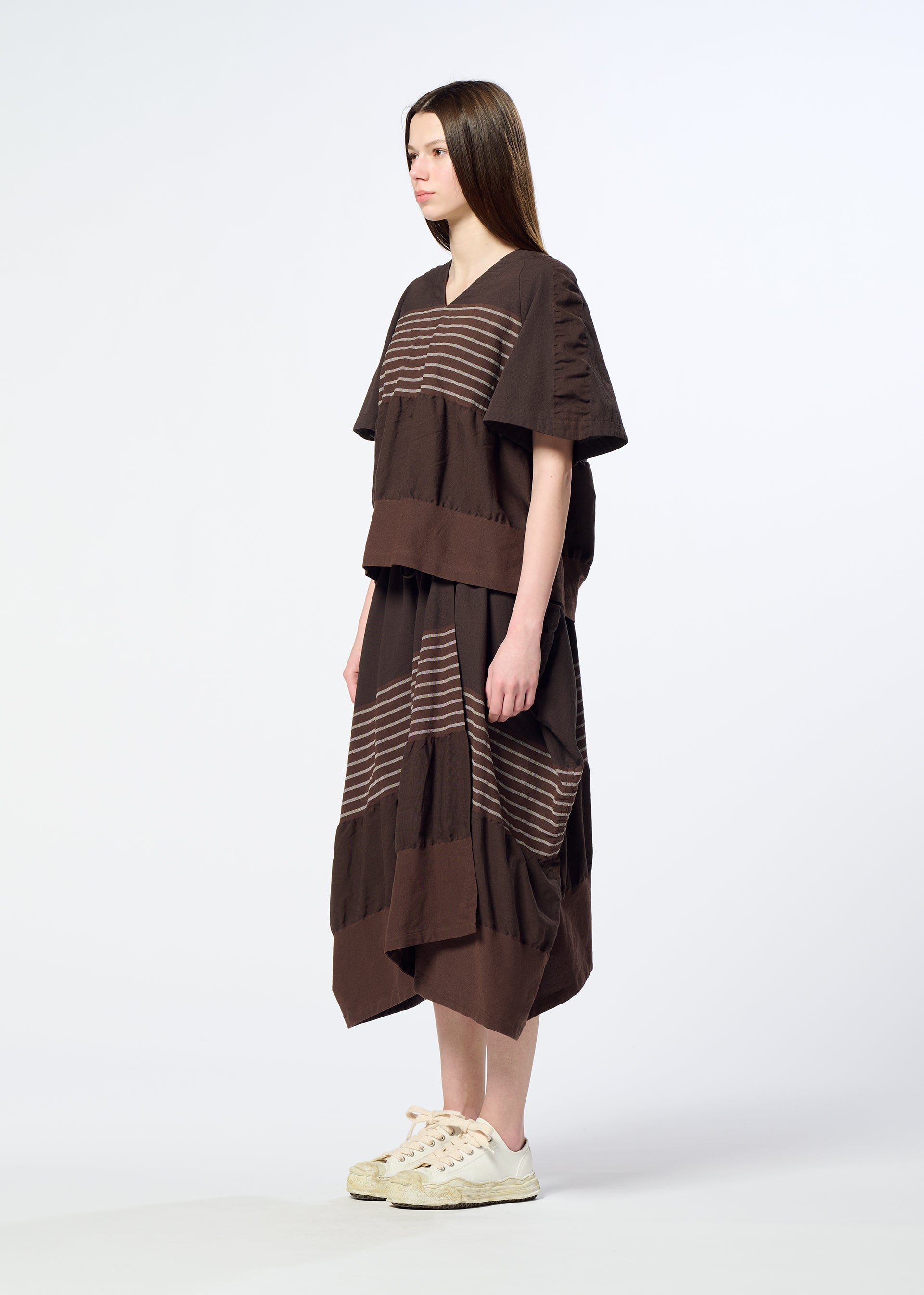 Zoomed view A woman stands in profile against a plain light background, wearing HaaT’s COLLAGE STRETCH Ⅱ—a relaxed, loose brown dress with horizontal beige stripes crafted from stretchy yarn—paired with white sneakers.