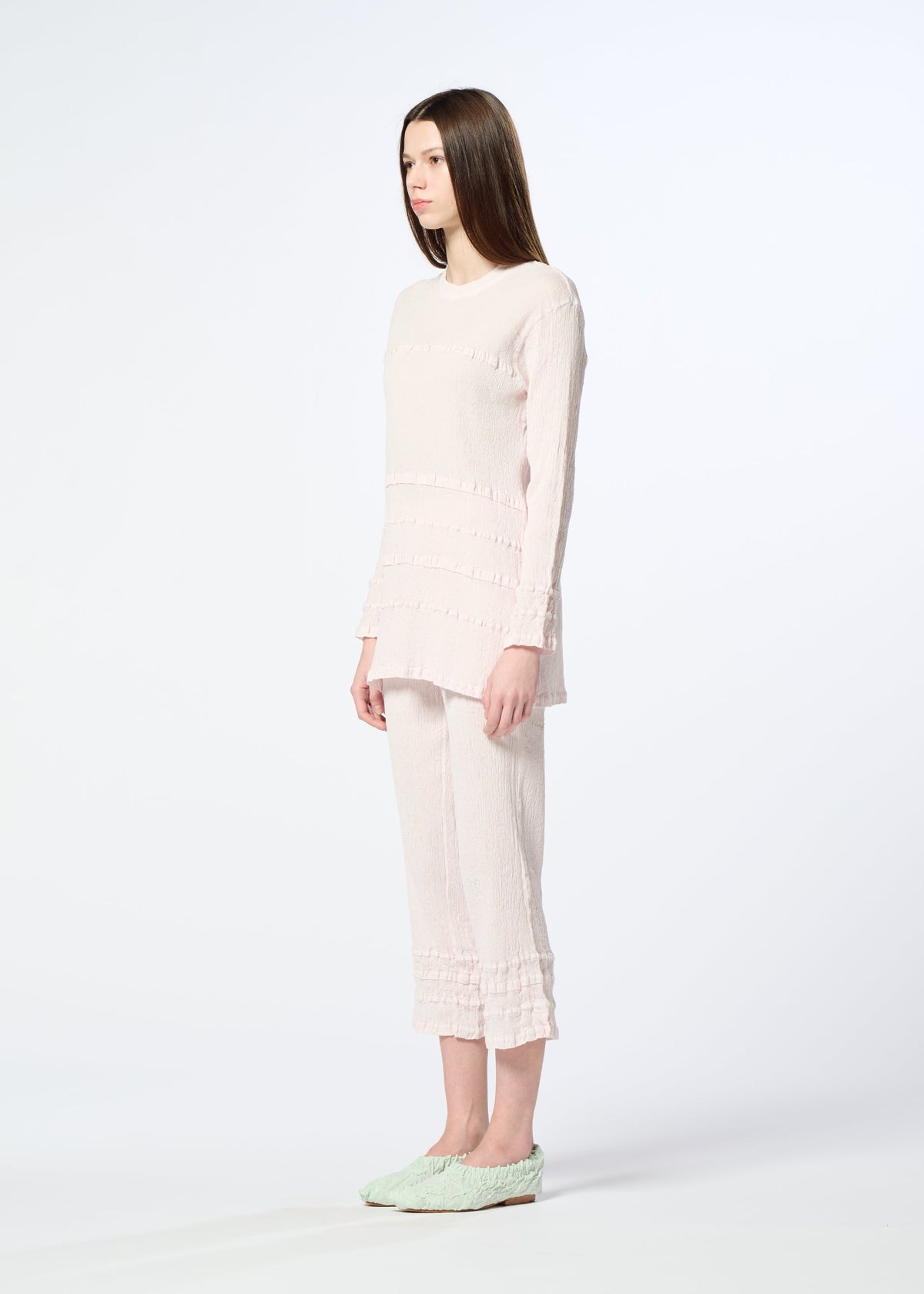 A woman with straight brown hair stands sideways in a long-sleeved, pale pink KYO CHIJIMI TSUCHIKABE set from HaaT, featuring subtle ruffles and a crepe-like texture, paired with light green slip-ons against a plain white background. view 7 of 8