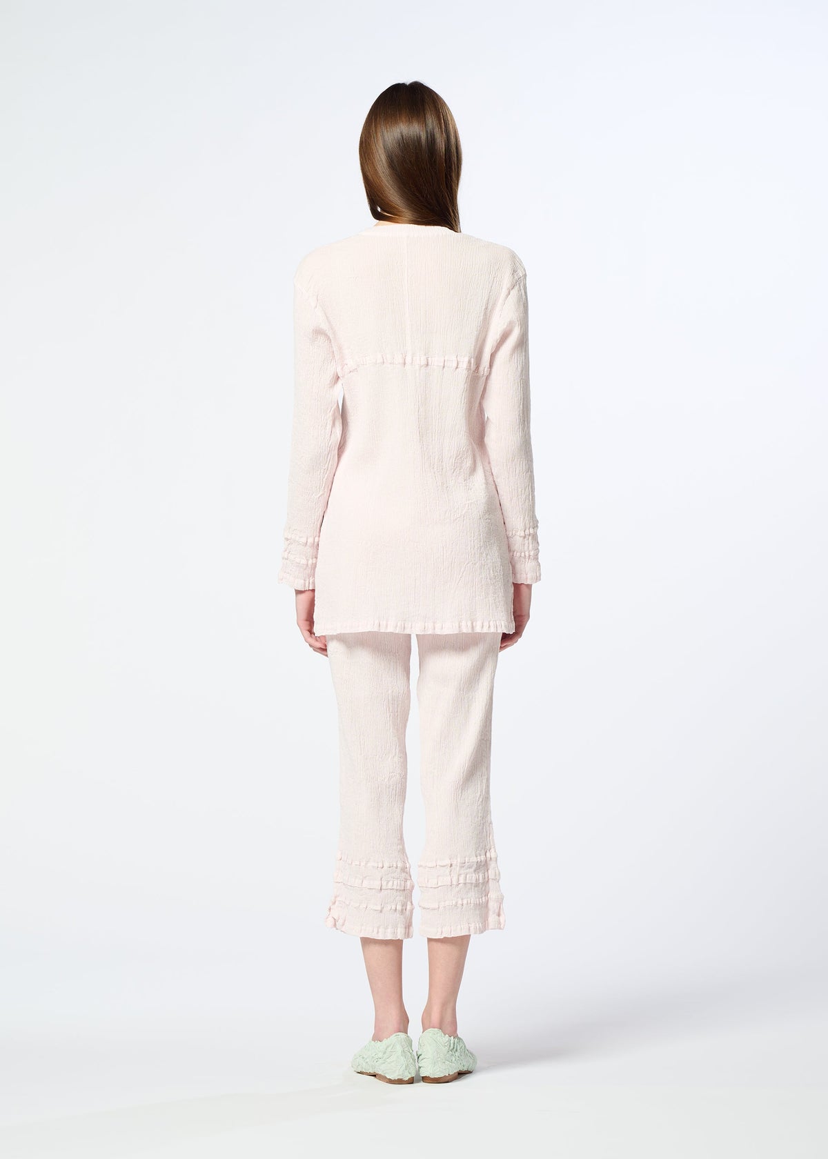 A woman faces away, wearing HaaT’s KYO CHIJIMI TSUCHIKABE light pink pajama set with a crepe-like texture and ruffle details, paired with mint green slippers, against a plain white background. view 8 of 8