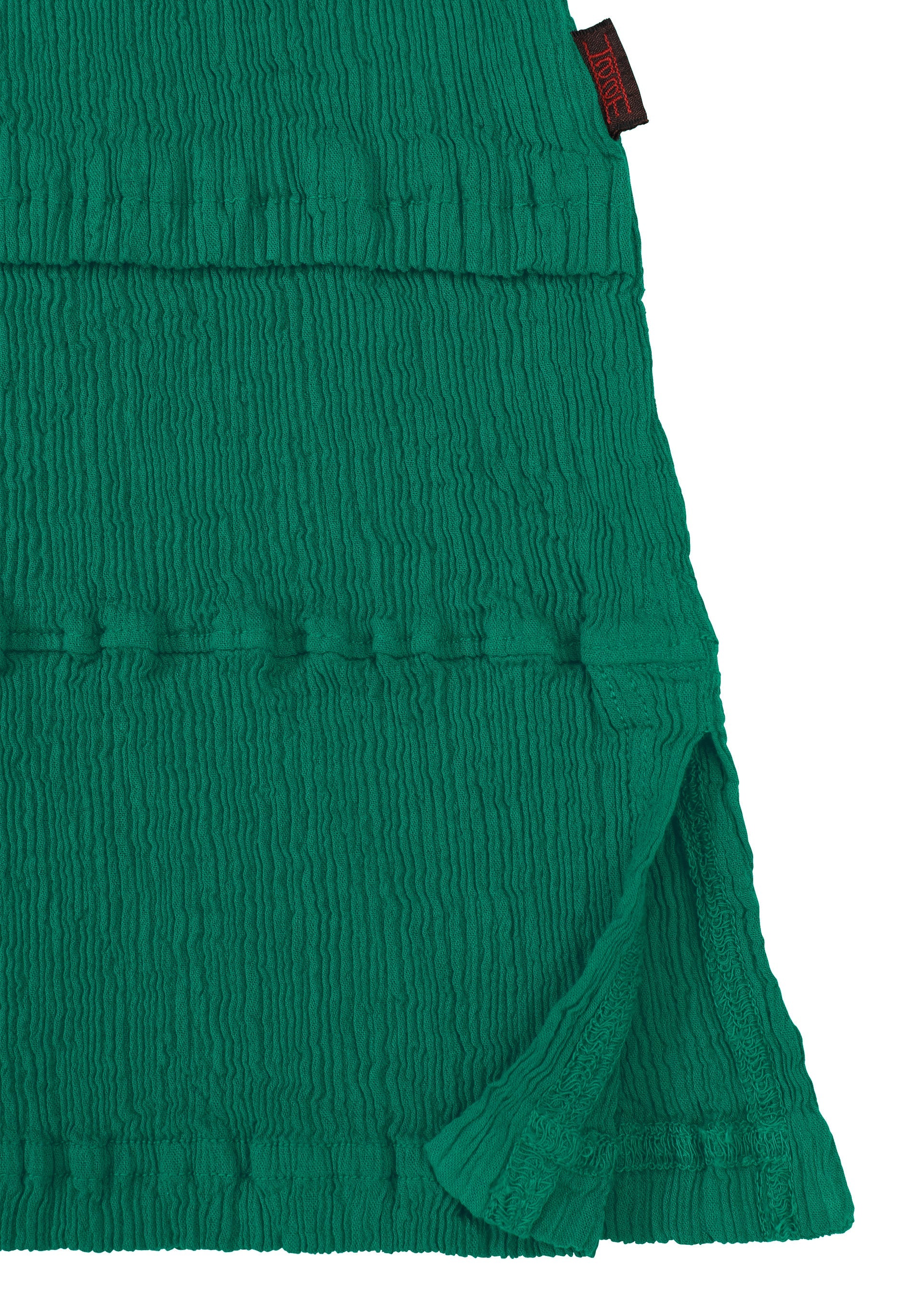 Zoomed view Close-up of the green KYO CHIJIMI TSUCHIKABE top by HaaT, featuring a crepe-like textured fabric with horizontal pleats, side slit, and a small black and red label at the top edge. Made from ultra-fine cotton.