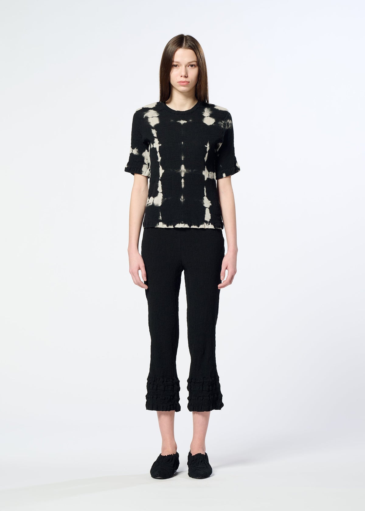 A woman with straight brown hair and a neutral expression wears HaaT’s KYO CHIJIMI PACKAGE DYE black and white crepe-like cotton top, styled with black cropped ruffled-hem pants and black shoes against a plain light background. view 6 of 8
