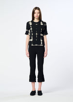A woman with straight brown hair and a neutral expression wears HaaT’s KYO CHIJIMI PACKAGE DYE black and white crepe-like cotton top, styled with black cropped ruffled-hem pants and black shoes against a plain light background.