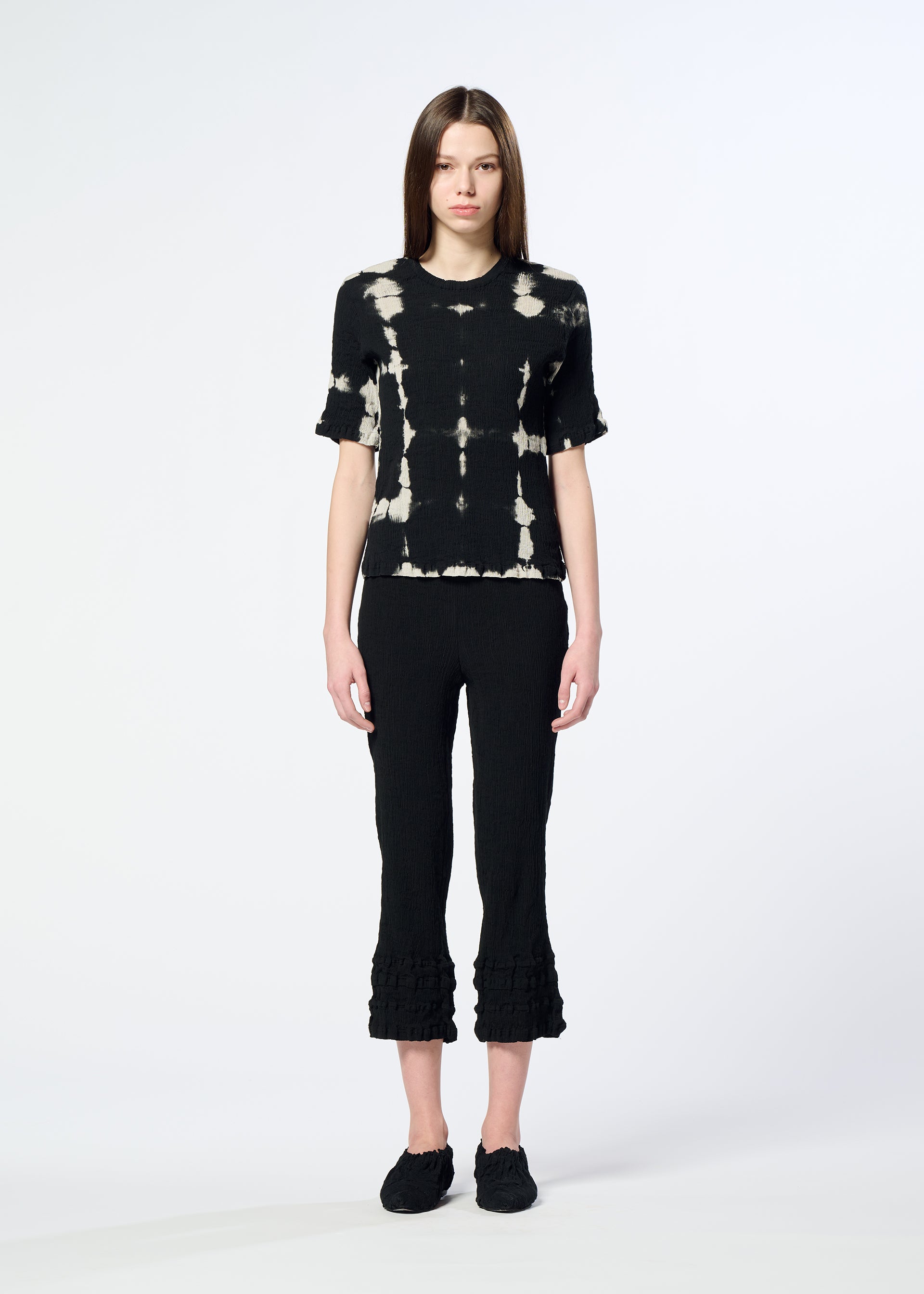 Zoomed view A woman with straight brown hair and a neutral expression wears HaaT’s KYO CHIJIMI PACKAGE DYE black and white crepe-like cotton top, styled with black cropped ruffled-hem pants and black shoes against a plain light background.