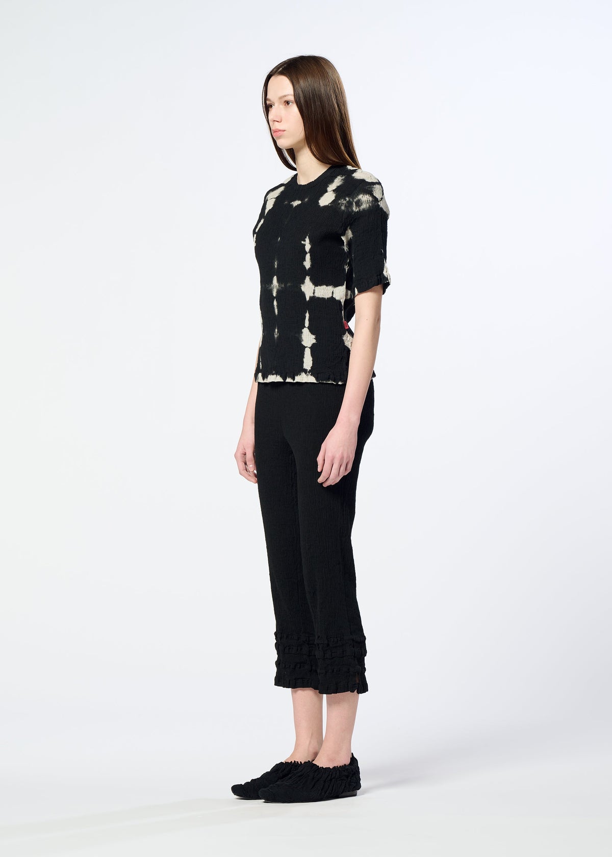 A woman with straight brown hair stands sideways, wearing HaaT's KYO CHIJIMI PACKAGE DYE top, black cropped pants with ruffled hems, and black slip-on shoes, showcasing Kyoto artisan craftsmanship against a light background. view 7 of 8