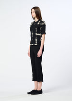 A woman with straight brown hair stands sideways, wearing HaaT's KYO CHIJIMI PACKAGE DYE top, black cropped pants with ruffled hems, and black slip-on shoes, showcasing Kyoto artisan craftsmanship against a light background.
