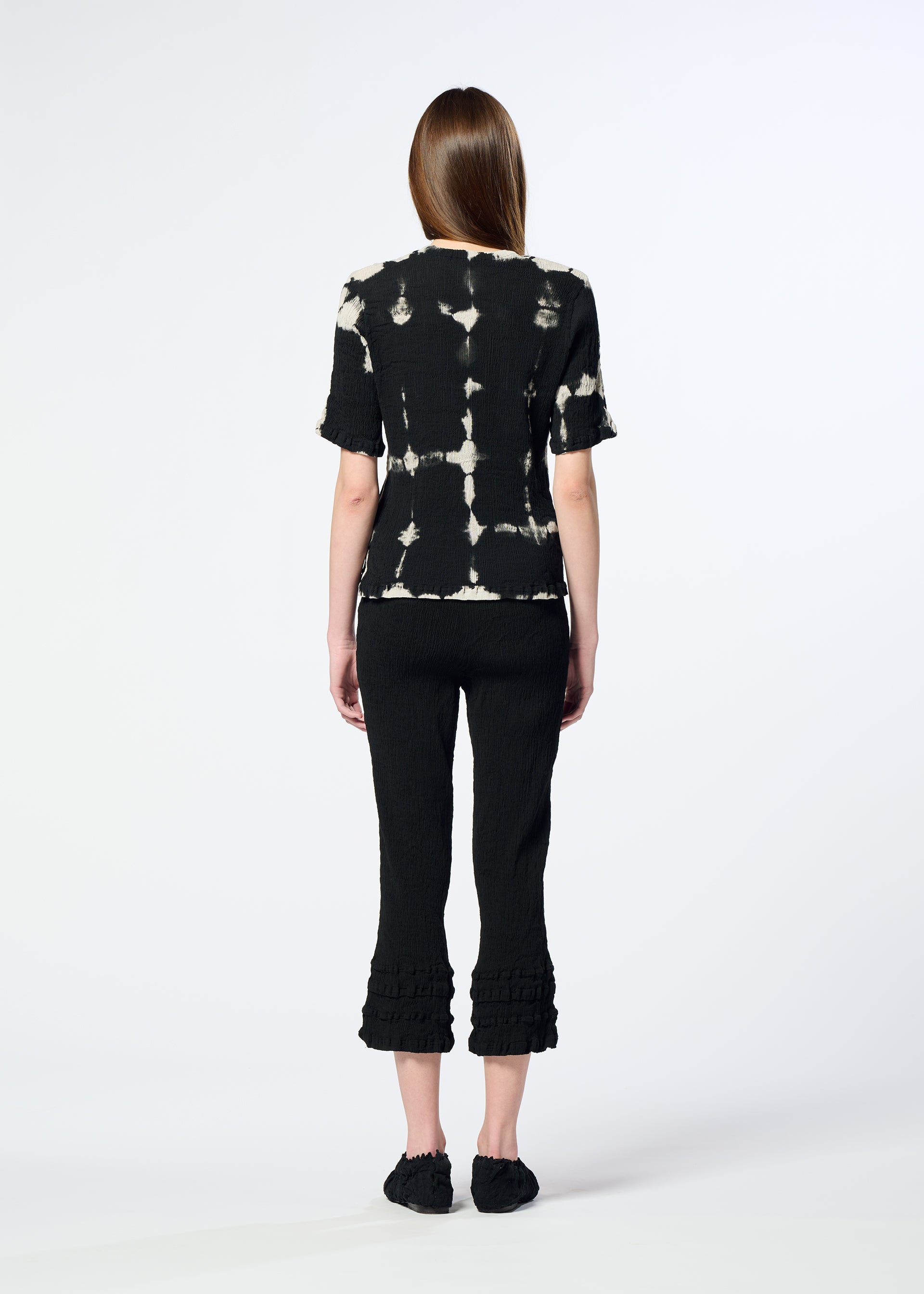 Zoomed view A person with long brown hair faces away, wearing a HaaT KYO CHIJIMI PACKAGE DYE black top with white tie-dye patterns, black cropped pants with ruffled hems, and black shoes, against a plain light background.