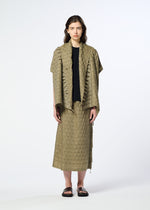 A woman stands facing forward against a plain white background, wearing HaaT’s HOLE & LINE outfit—an olive-green textured skirt with eyelet embroidery and a matching short-sleeve jacket over a black shirt, paired with black sandals.