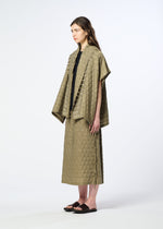 A woman stands sideways against a plain light background, wearing HaaT’s HOLE & LINE textured olive-green cape and matching long skirt with delicate eyelet embroidery, paired with a black top and black sandals.