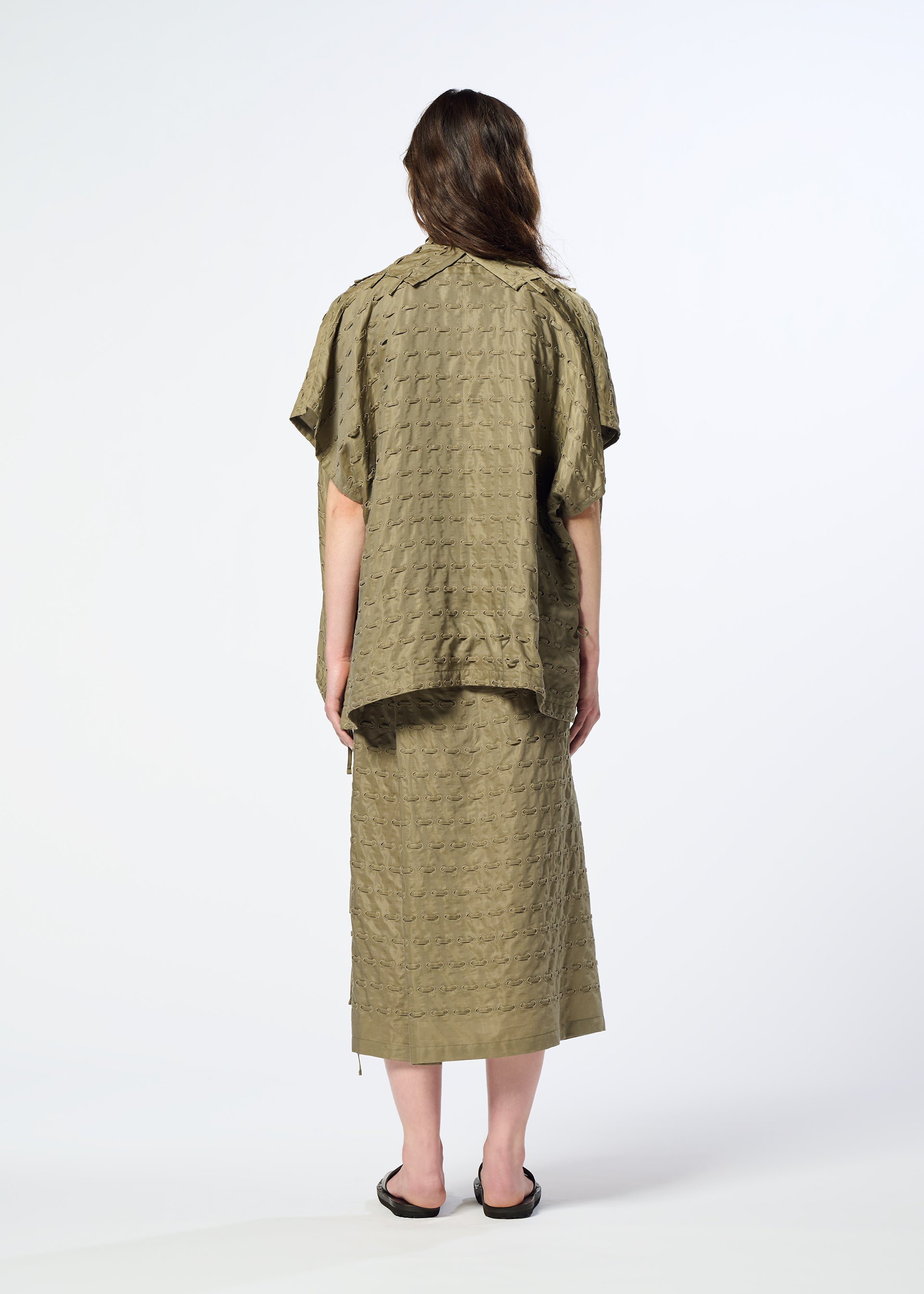 Zoomed view A person stands facing away, wearing the HaaT HOLE & LINE olive textured short-sleeve top with subtle eyelet embroidery and the matching mid-length skirt, paired with black sandals. The background is plain white.