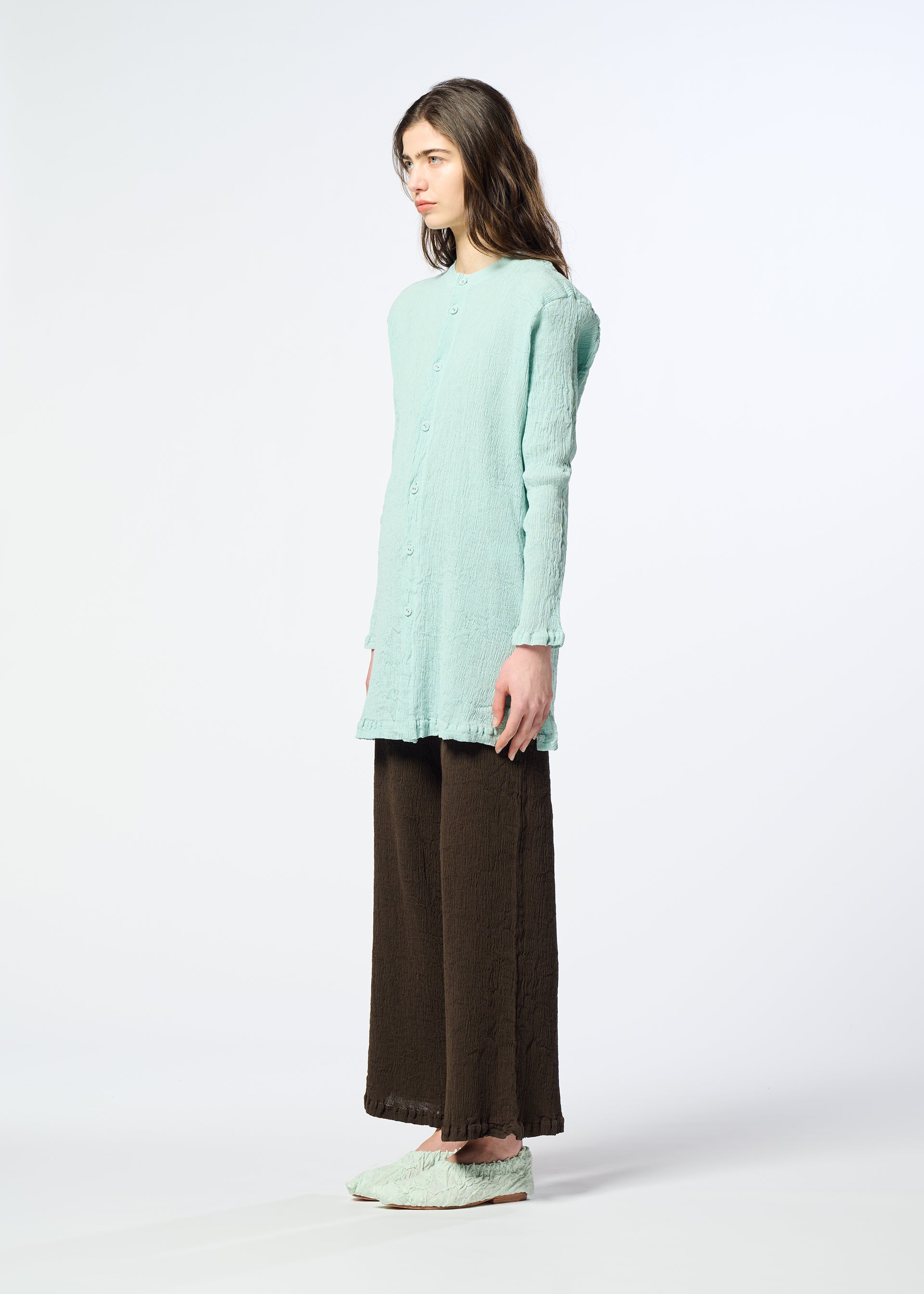 Zoomed view A person stands sideways against a plain white background, wearing the HaaT KYO CHIJIMI AUGUST textured light blue long-sleeved cardigan, loose brown pants, and matching light blue shoes.