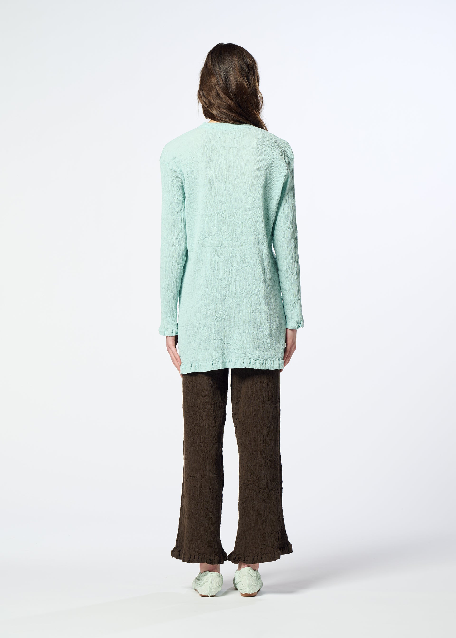Zoomed view A person stands facing away, wearing the HaaT KYO CHIJIMI AUGUST long light blue cardigan with a crinkled texture, paired with brown textured pants and light-colored shoes. The background is plain white.