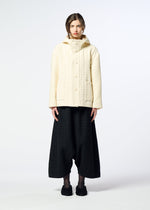 A person stands facing forward in a cream SAKIORI hooded jacket from HaaT and loose black pants against a plain white background, showcasing minimalist, contemporary style with traditional weaving influences.