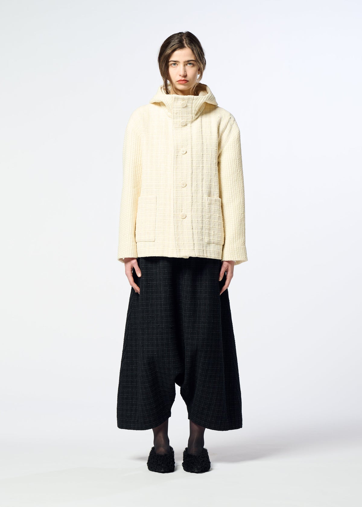Zoomed view A person stands facing forward in a cream SAKIORI hooded jacket from HaaT and loose black pants against a plain white background, showcasing minimalist, contemporary style with traditional weaving influences.