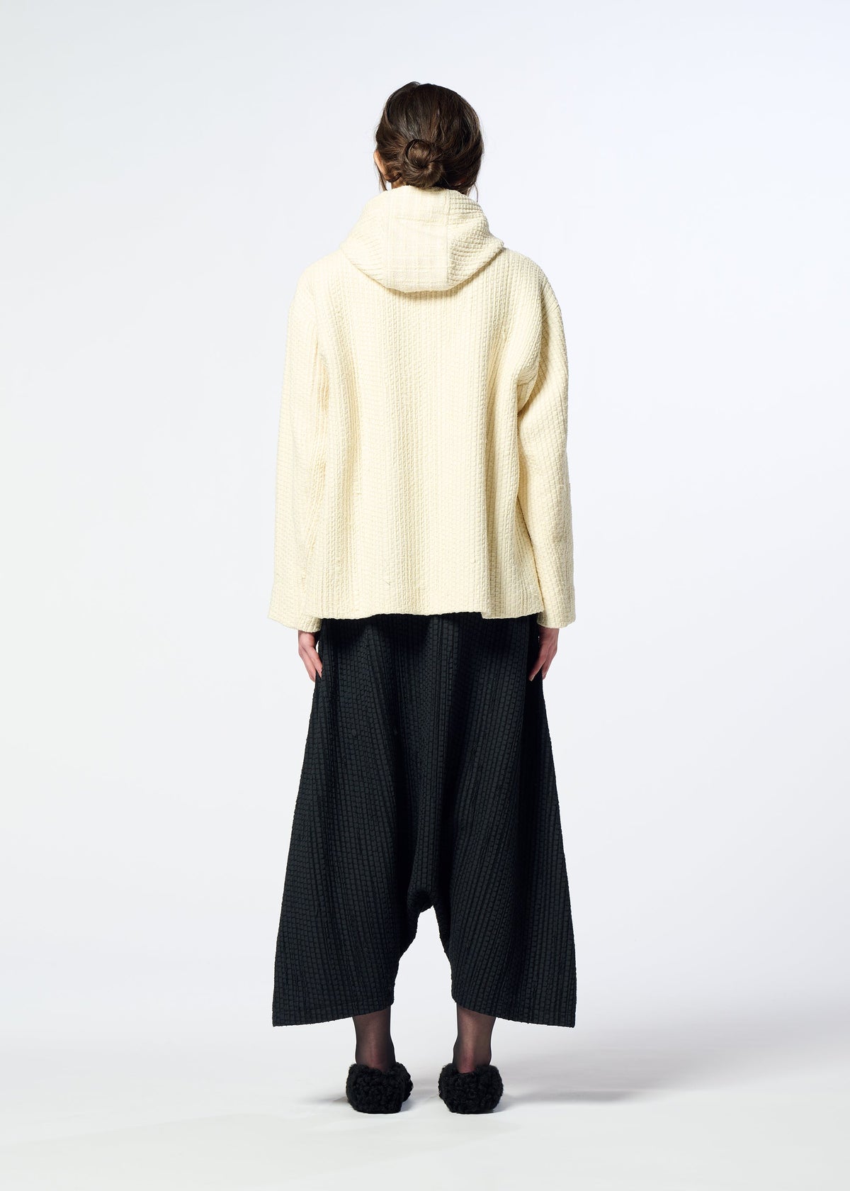 A person stands facing away, wearing the cream-colored HaaT SAKIORI hooded jacket, wide black textured pants, and black slippers against a plain white background. view 10 of 10