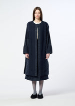 A woman stands facing forward against a plain light background, wearing HaaT’s COLLAGE STRETCH Ⅰ—a long, dark blue textured coat with a subtle large check pattern—layered over a matching skirt, paired with gray tights and black shoes.