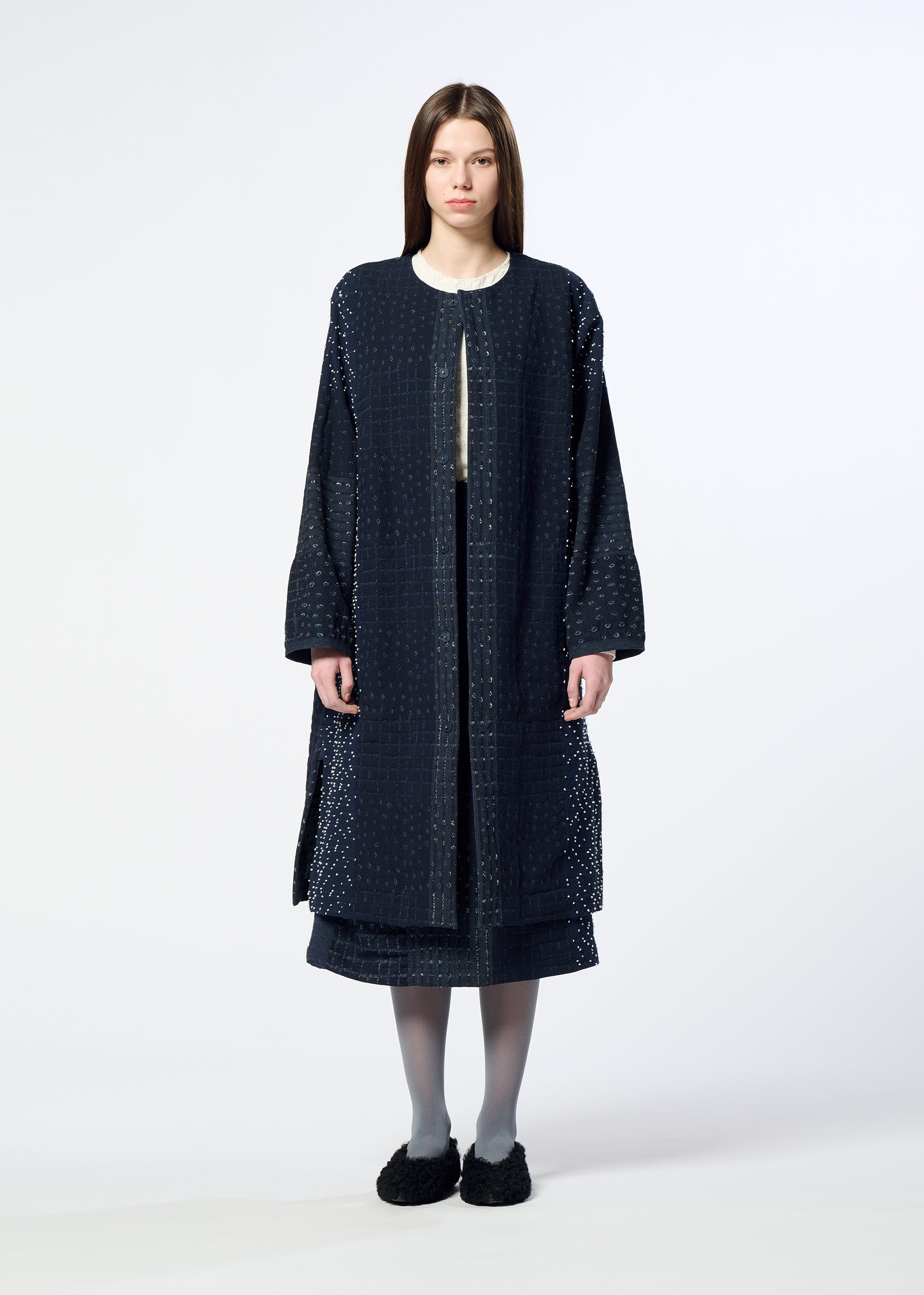 Zoomed view A woman stands facing forward against a plain light background, wearing HaaT’s COLLAGE STRETCH Ⅰ—a long, dark blue textured coat with a subtle large check pattern—layered over a matching skirt, paired with gray tights and black shoes.