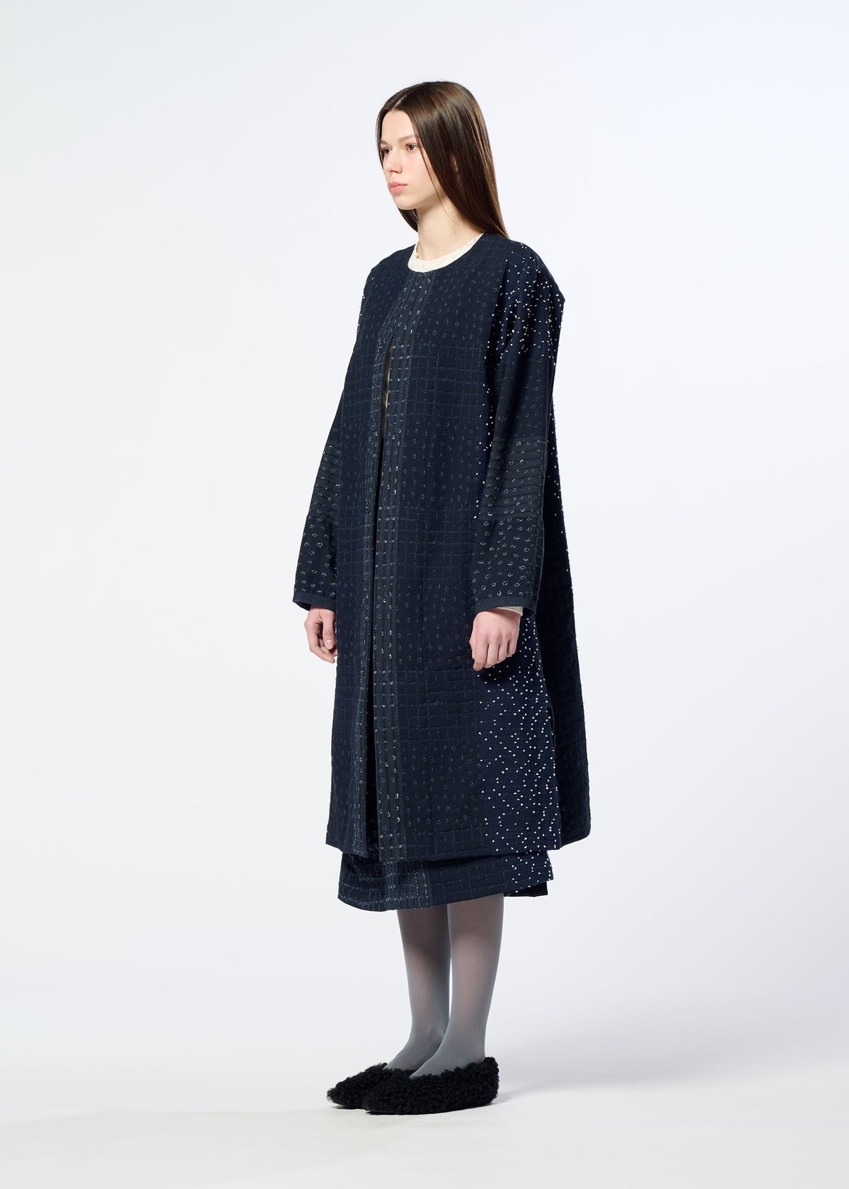 A woman with straight brown hair wears HaaT’s COLLAGE STRETCH Ⅰ—a long, loose-fitting navy blue coat with a subtle large check pattern—paired with a matching skirt, gray tights, and black slip-on shoes, standing against a plain white background. view 7 of 8