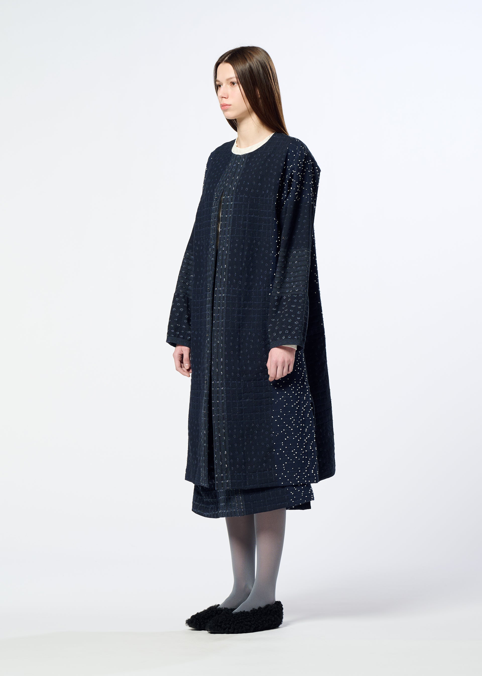 Zoomed view A woman with straight brown hair wears HaaT’s COLLAGE STRETCH Ⅰ—a long, loose-fitting navy blue coat with a subtle large check pattern—paired with a matching skirt, gray tights, and black slip-on shoes, standing against a plain white background.
