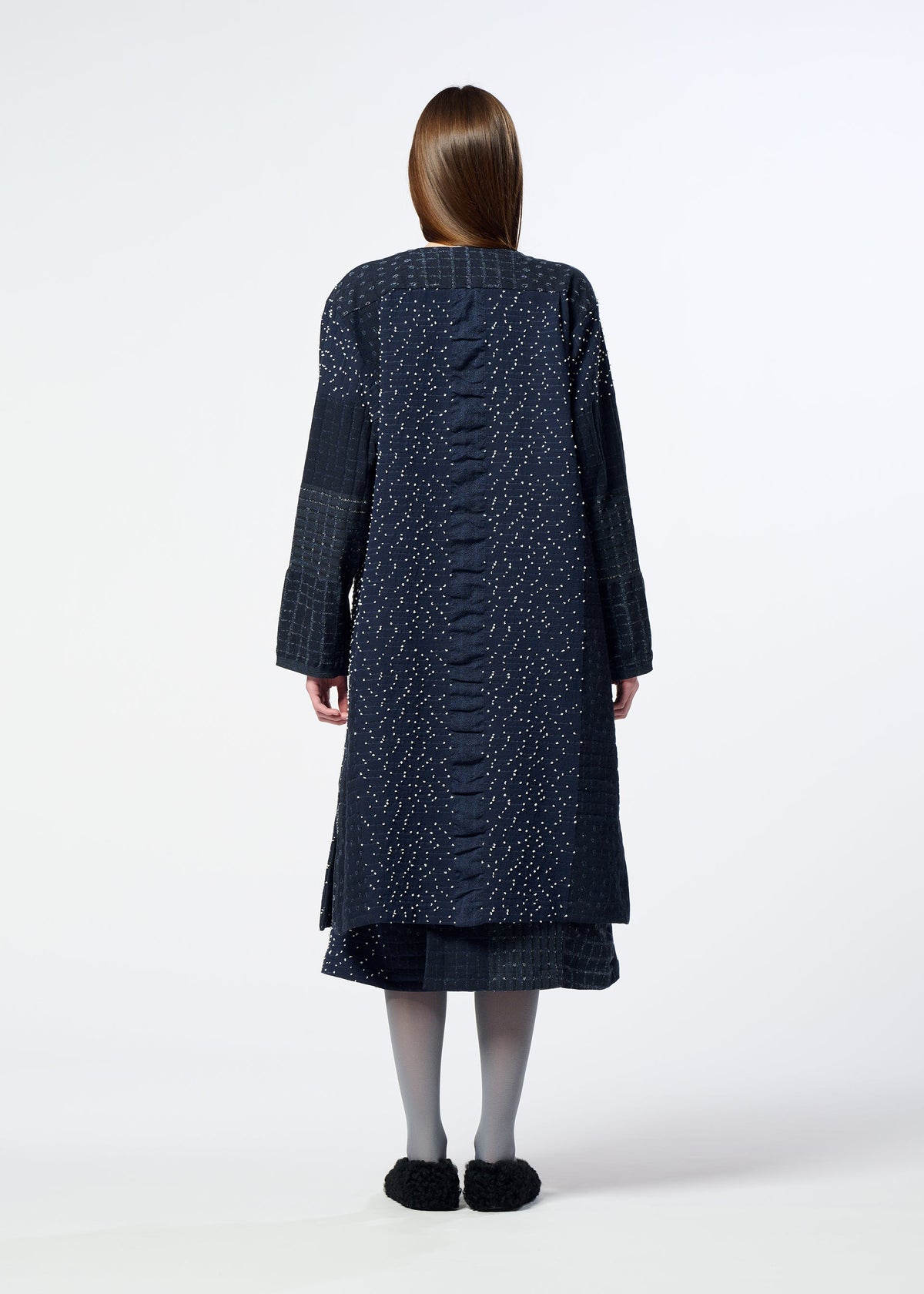 A person with long hair stands facing away, wearing the HaaT COLLAGE STRETCH Ⅰ knee-length coat featuring a dark, large check pattern, styled with a skirt, gray tights, and black shoes against a plain white background. view 8 of 8