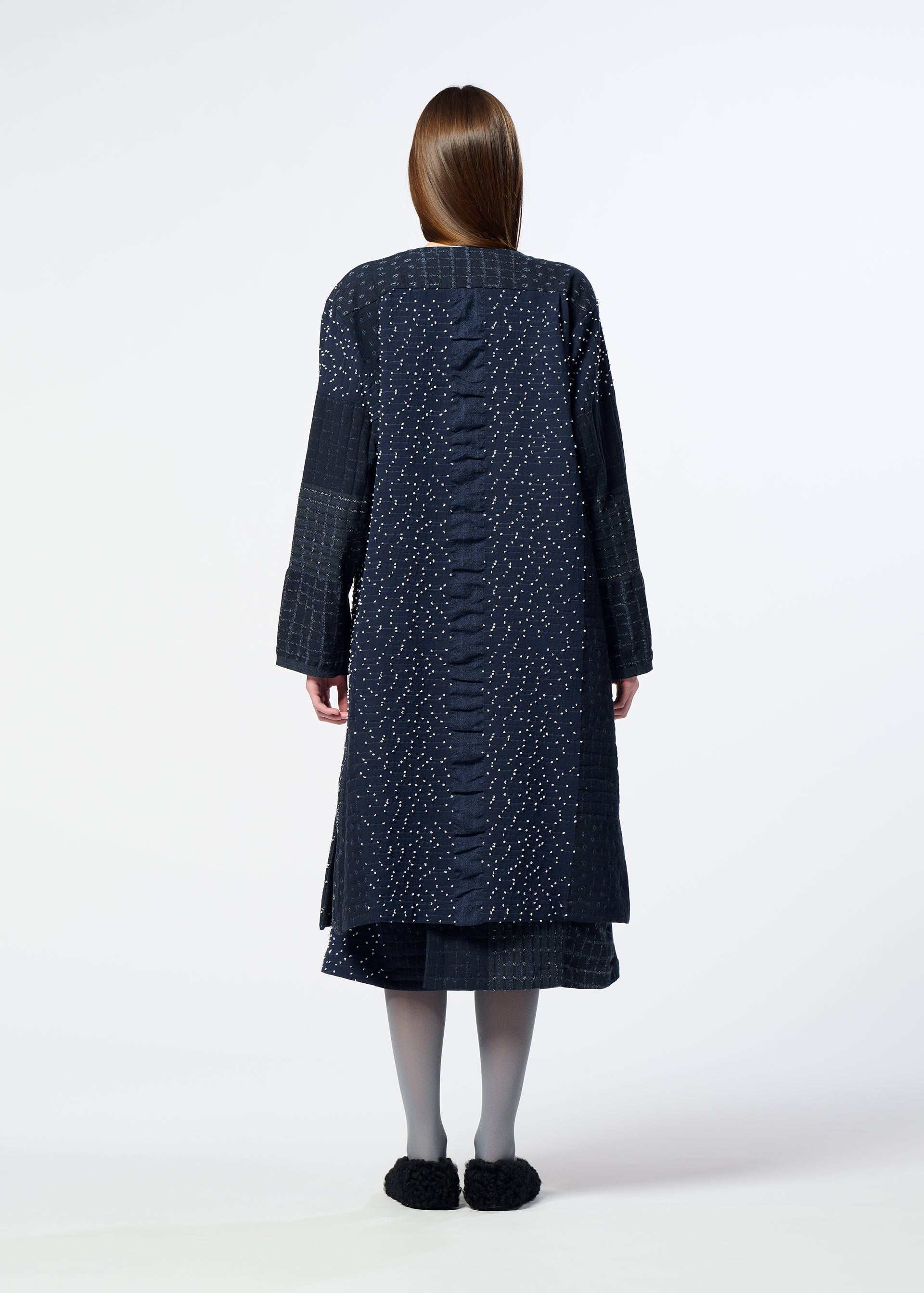 Zoomed view A person with long hair stands facing away, wearing the HaaT COLLAGE STRETCH Ⅰ knee-length coat featuring a dark, large check pattern, styled with a skirt, gray tights, and black shoes against a plain white background.