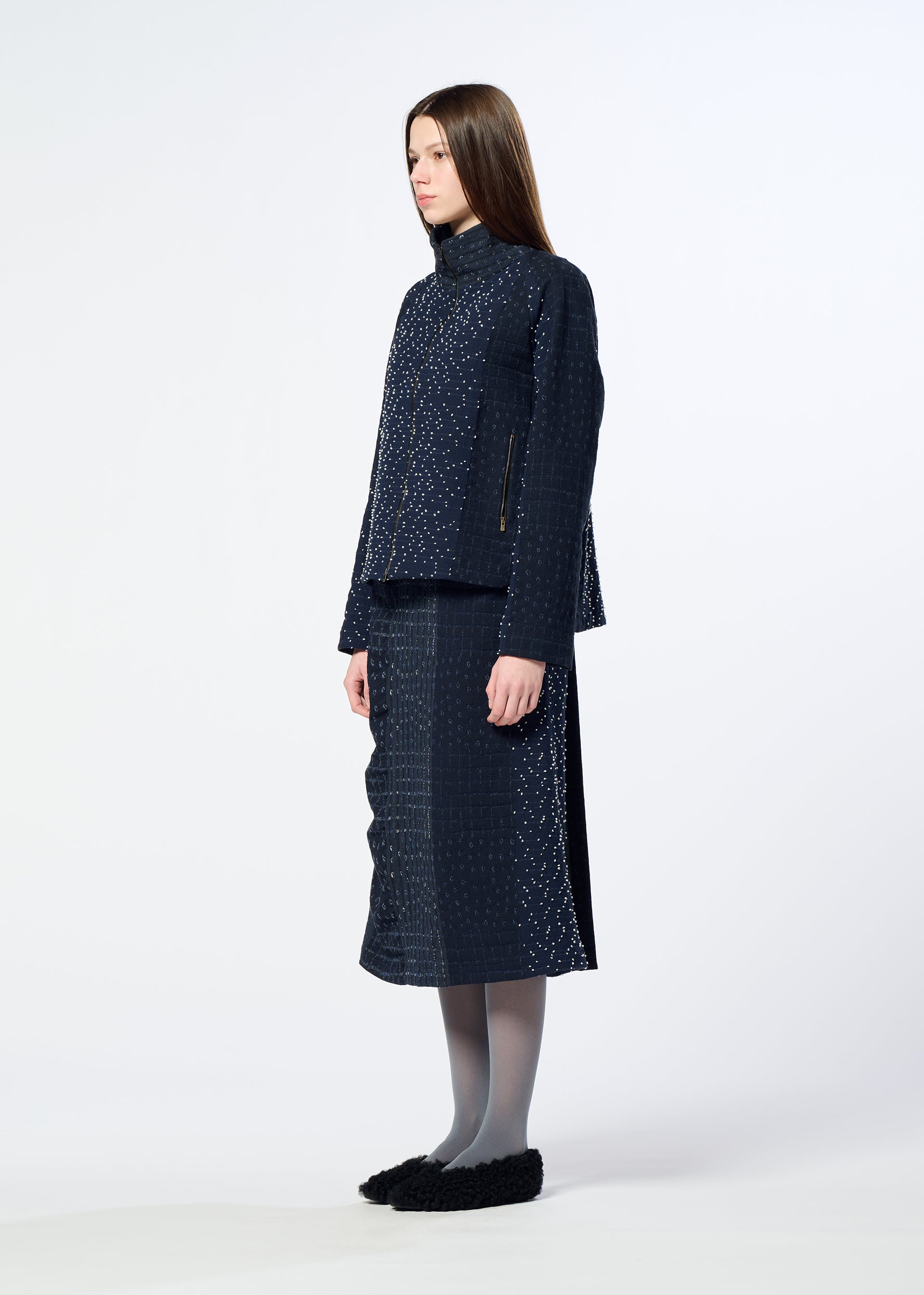 Zoomed view A woman stands in profile against a plain light background wearing HaaT’s COLLAGE STRETCH Ⅰ navy check pattern jacket and matching midi skirt, paired with gray tights and black shoes.