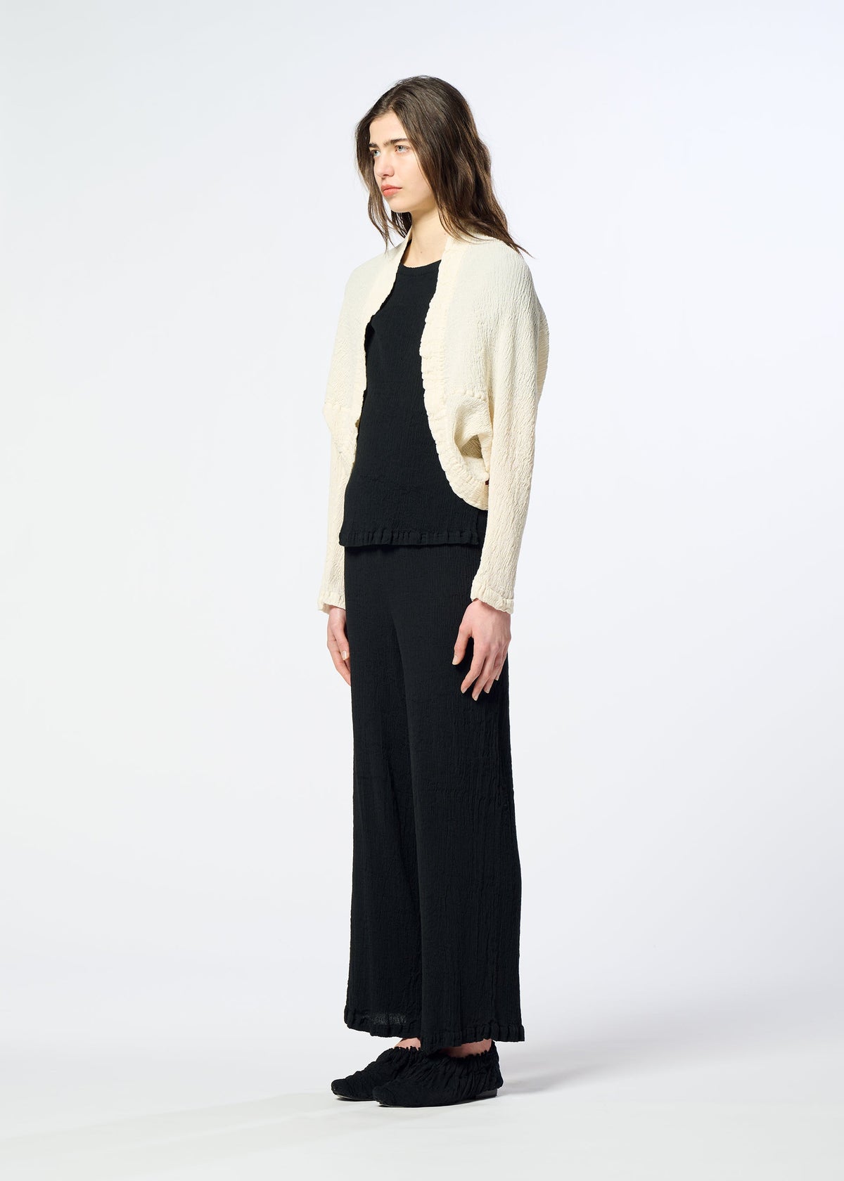 A person stands sideways against a plain white background, wearing HaaT’s KYO CHIJIMI 2WAY wide-leg pants and a black top, paired with a textured, cream-colored bolero-style cardigan. Their long brown hair and black shoes complement the look, highlighting the subtle crinkle texture inspired by KYO CHIJIMI. view 7 of 8