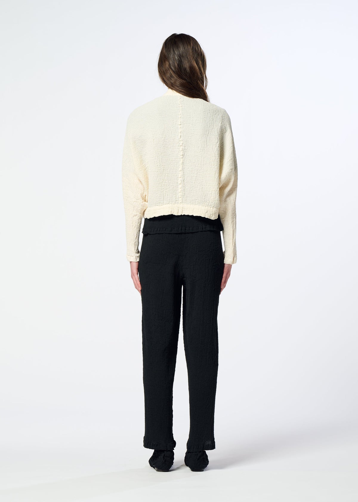 A person with long brown hair, seen from behind, wears a cream-colored HaaT KYO CHIJIMI 2WAY bolero-style cardigan with a subtle crinkle texture and black pants. They stand on a white background with their arms relaxed by their sides. view 8 of 8
