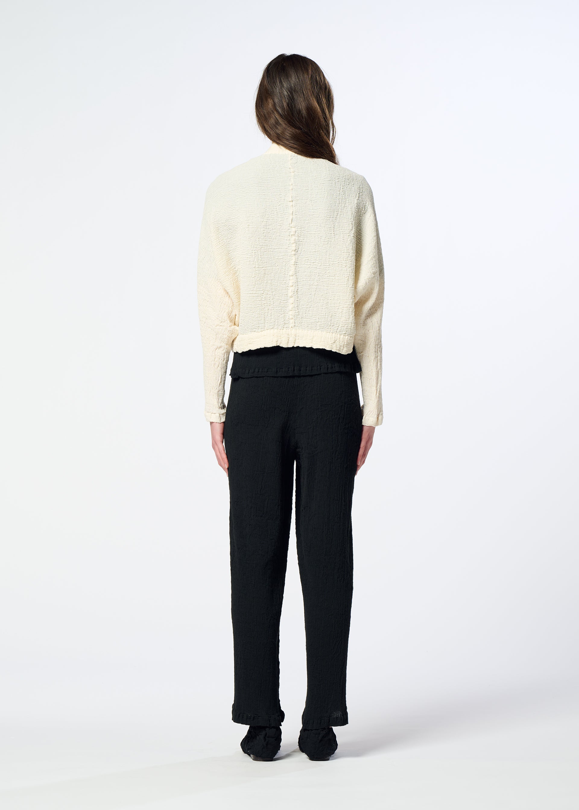 Zoomed view A person with long brown hair, seen from behind, wears a cream-colored HaaT KYO CHIJIMI 2WAY bolero-style cardigan with a subtle crinkle texture and black pants. They stand on a white background with their arms relaxed by their sides.