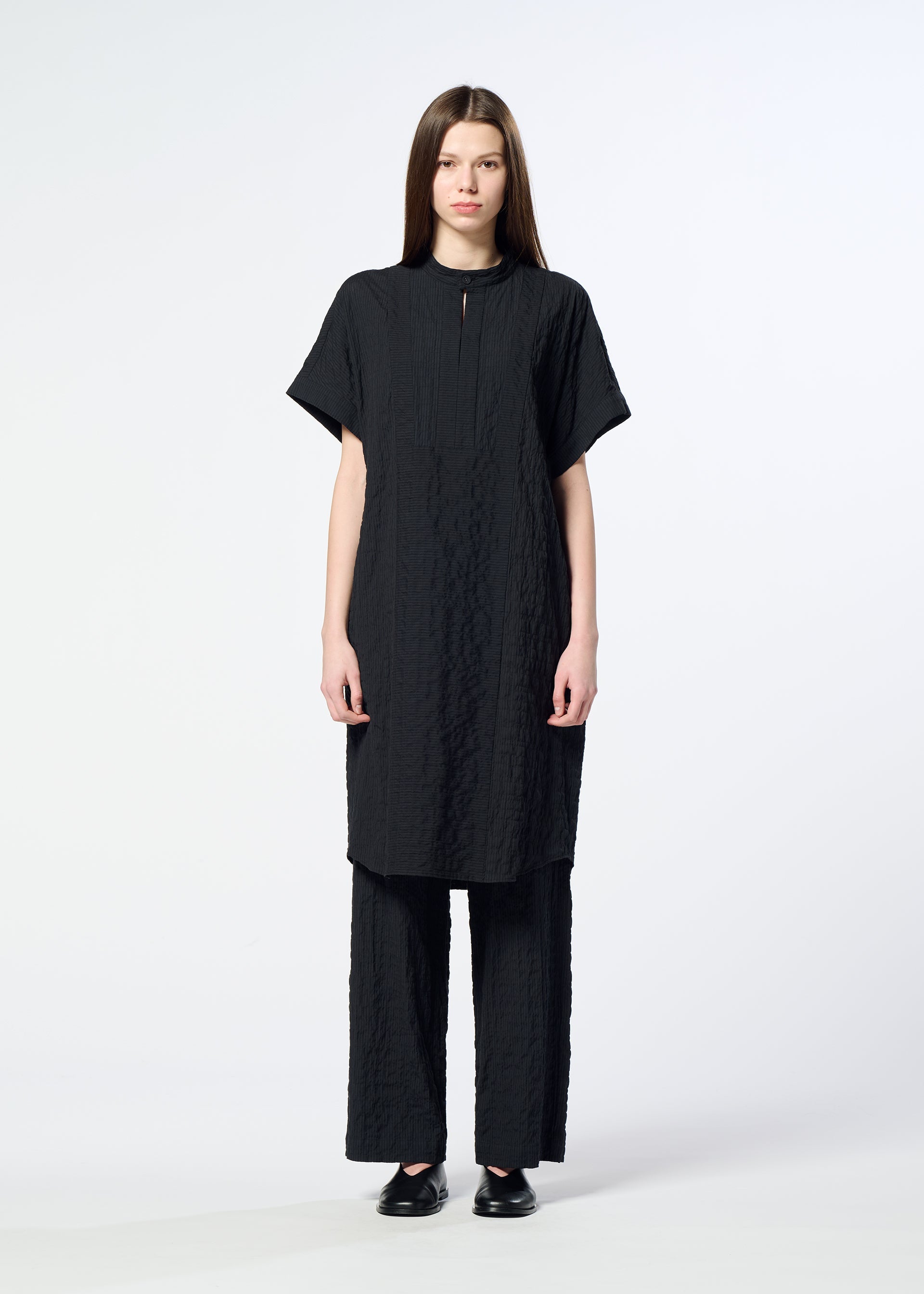Zoomed view A woman stands facing forward against a plain white background, wearing HaaT’s RIPPLE STRIPE textured black knee-length seersucker tunic over matching wide-leg pants and black shoes.