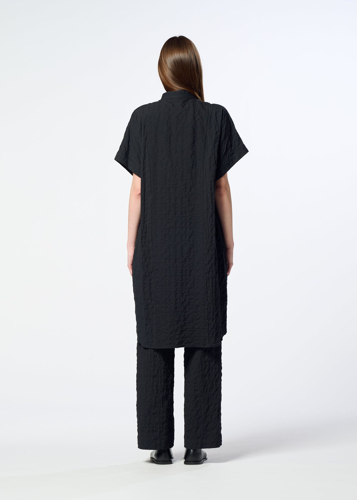 A person stands facing away, dressed in the HaaT RIPPLE STRIPE textured short-sleeved tunic and matching loose black pants, both featuring a subtle wave stripe pattern, set against a plain white background. view 8 of 8
