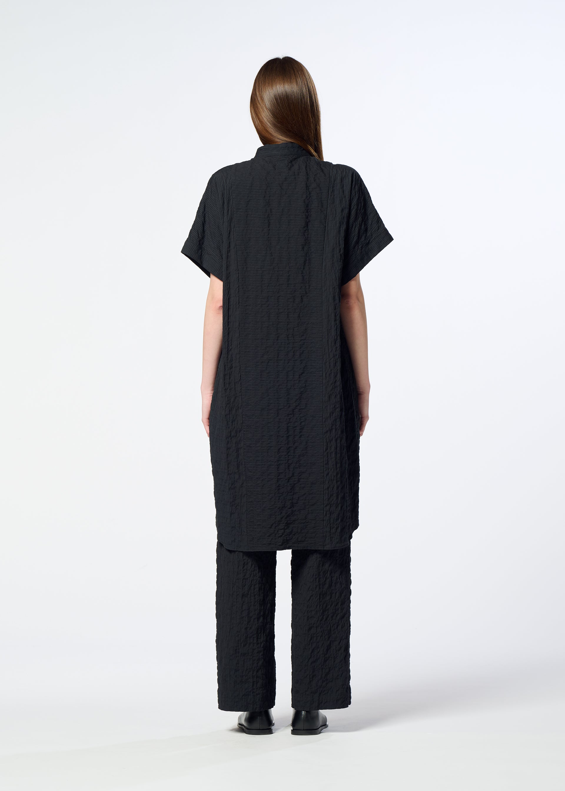 Zoomed view A person stands facing away, dressed in the HaaT RIPPLE STRIPE textured short-sleeved tunic and matching loose black pants, both featuring a subtle wave stripe pattern, set against a plain white background.