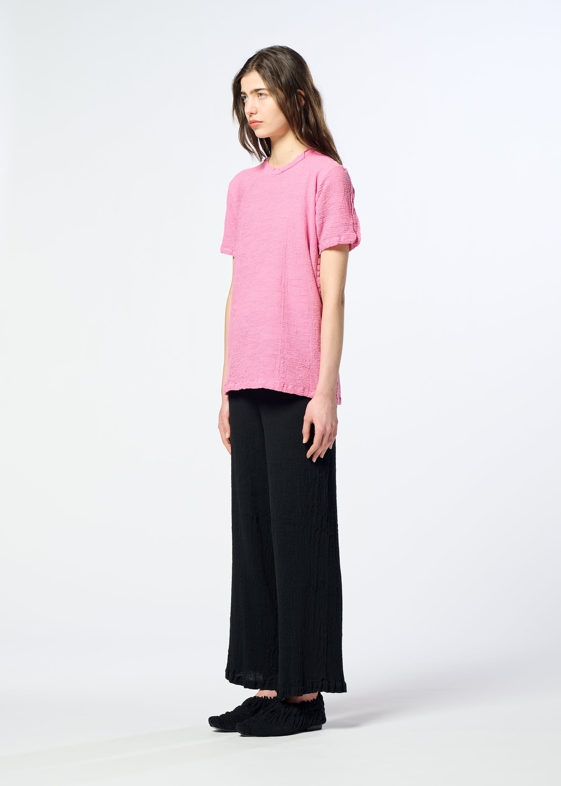 Zoomed view A person with long hair stands sideways against a white background, wearing the HaaT KYO CHIJIMI 2WAY short-sleeved tunic in pink, crafted using garment dyeing techniques, paired with black wide-leg pants and black shoes.