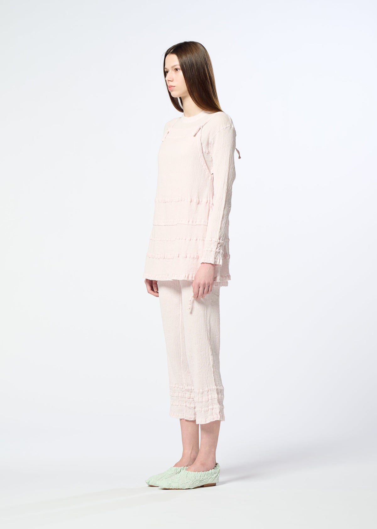 A woman with long straight hair stands in profile against a white background, wearing HaaT’s KYO CHIJIMI TSUCHIKABE apron-style top and matching ruffle-detailed cropped pants, crafted from ultra-fine cotton, paired with light green slip-on shoes. view 8 of 10