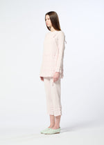 A woman with long straight hair stands in profile against a white background, wearing HaaT’s KYO CHIJIMI TSUCHIKABE apron-style top and matching ruffle-detailed cropped pants, crafted from ultra-fine cotton, paired with light green slip-on shoes.