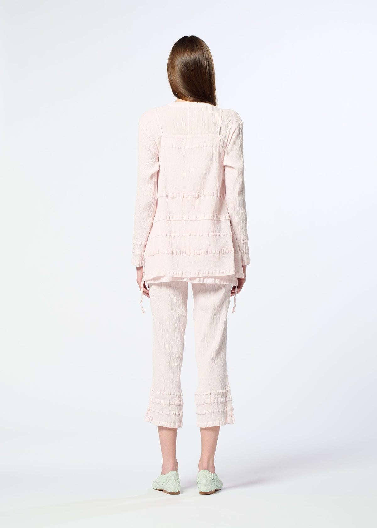 A woman with long brown hair faces away, wearing HaaT’s KYO CHIJIMI TSUCHIKABE apron-style top and matching ruffled cropped pants with a crepe texture in light pink, plus mint green shoes, all set against a plain white background. view 9 of 10