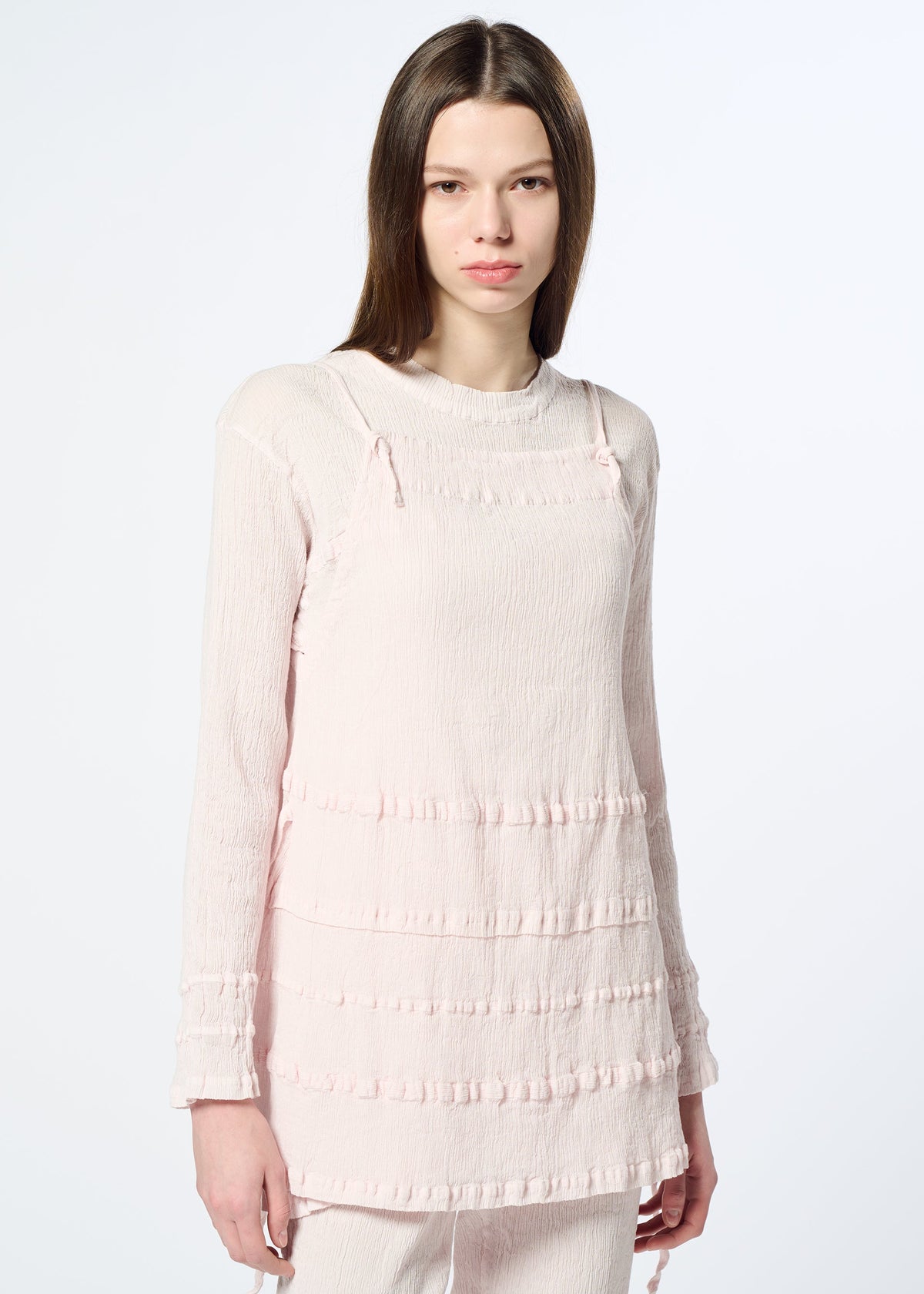 A young woman with straight brown hair wears HaaT’s KYO CHIJIMI TSUCHIKABE light pink apron-style top and matching pants with textured stripes and a crepe-like cotton feel, standing against a plain white background. view 10 of 10