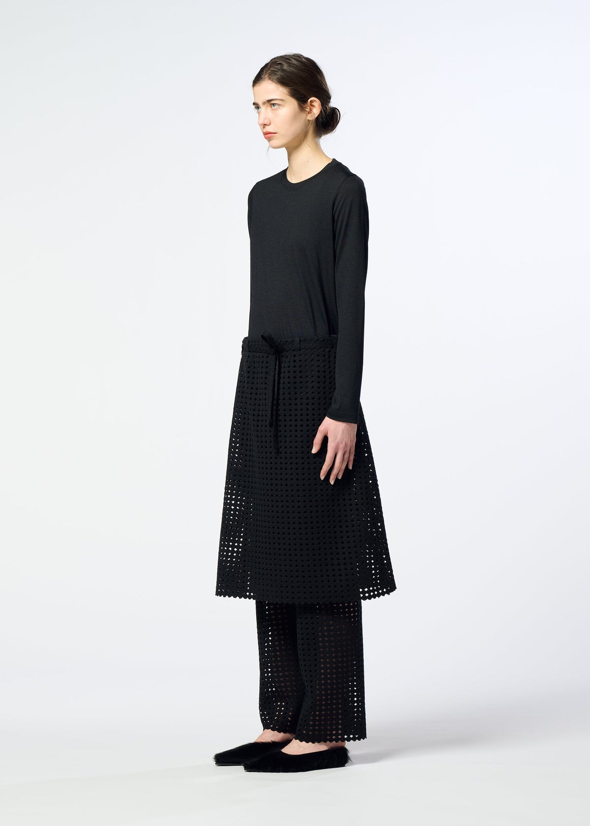 Zoomed view A person stands sideways against a plain light background, wearing HaaT’s LACY UNDER long-sleeved black top, a black perforated skirt over ponte jersey stretch pants, and black shoes.