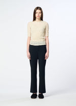 A person with long brown hair stands against a plain white background, wearing HaaT's PONTE JERSEY short-sleeved textured cream top, flowy pants for relaxed comfort, and black shoes.