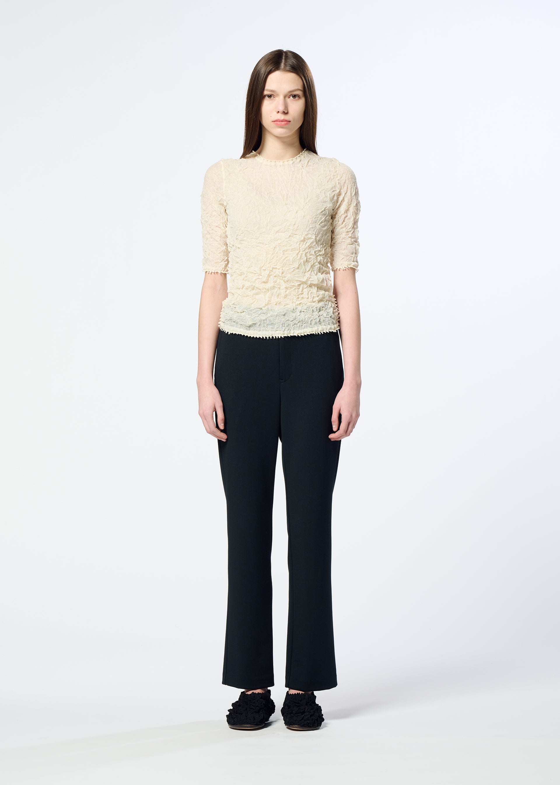 Zoomed view A person with long brown hair stands against a plain white background, wearing HaaT's PONTE JERSEY short-sleeved textured cream top, flowy pants for relaxed comfort, and black shoes.